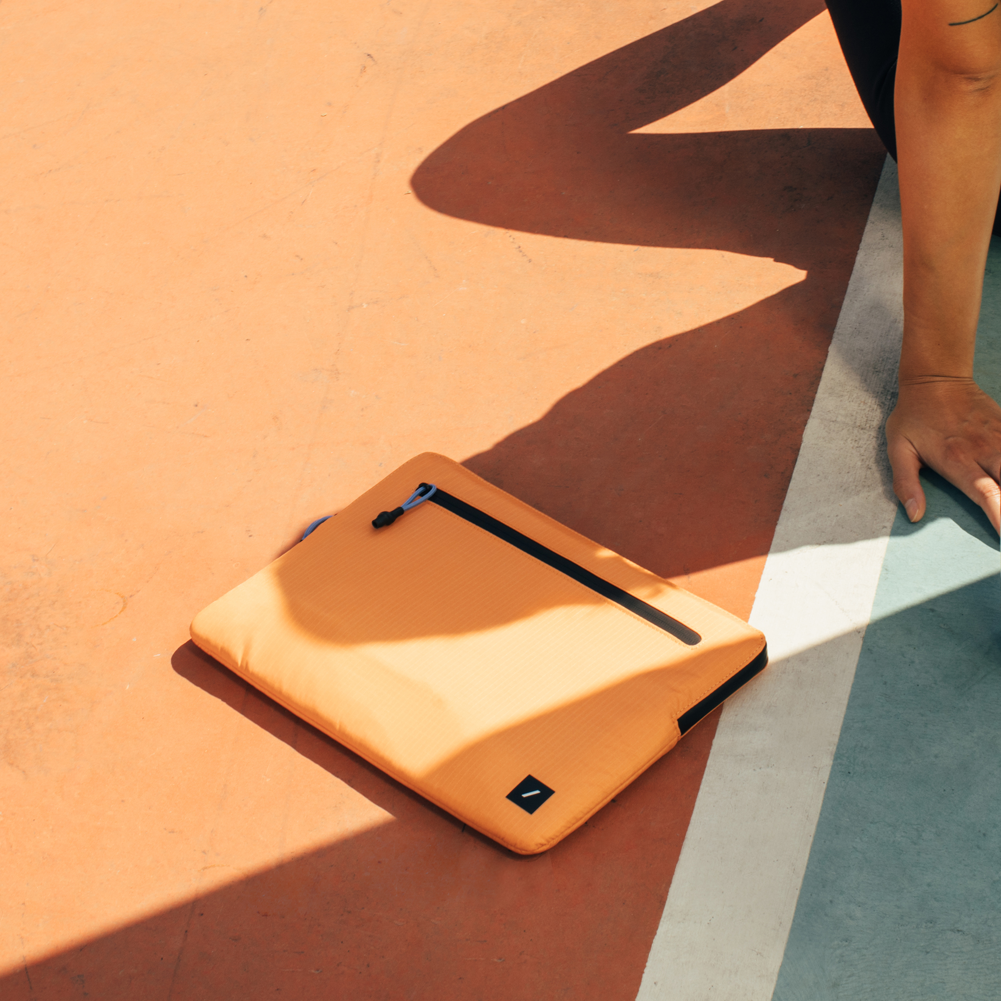 Orangene Laptoptasche auf einer orangefarbenen und blauen Oberfläche. Arm einer Person ruht daneben.