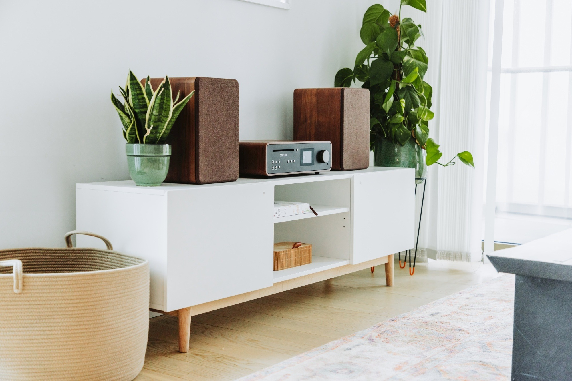 Mobile bianco con altoparlanti stereo, uno stereo e piante verdi sopra. Cesto beige sul pavimento.