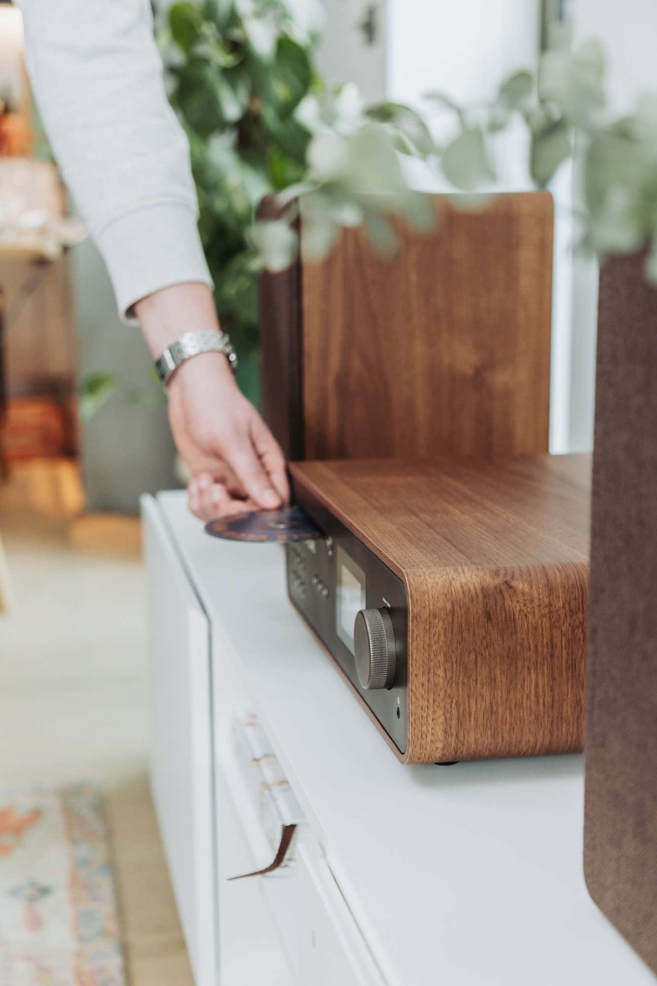 Una persona inserisce un CD in un sistema audio in legno su un mobile bianco. Piante sullo sfondo.