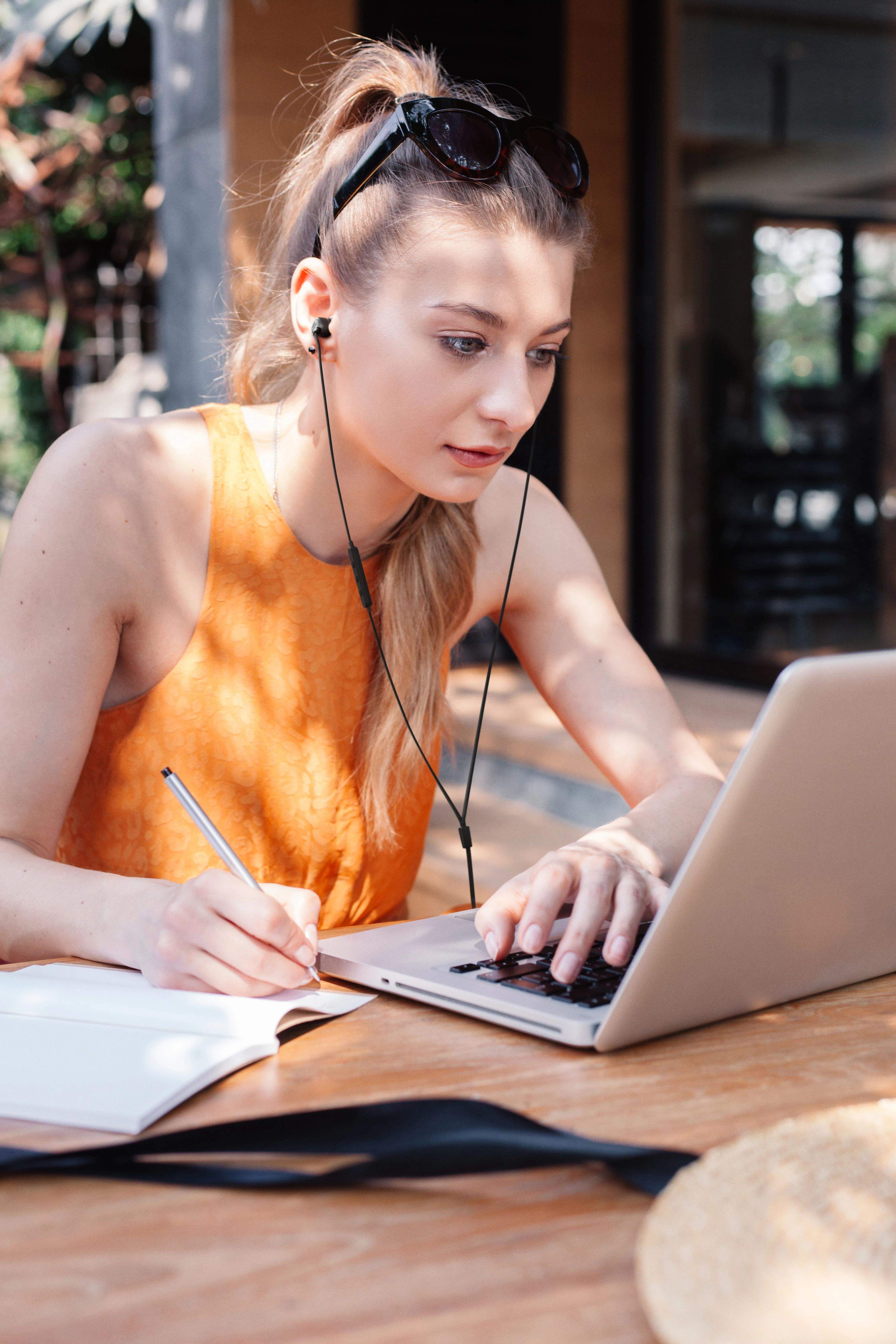 Turuncu üstlü kadın, laptop kullanıyor, not defterine yazıyor, güneş gözlüğü takılı.