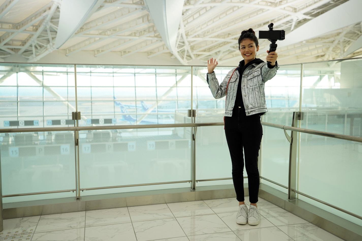 Una mujer con chaqueta plateada se filma con un teléfono en un aeropuerto.