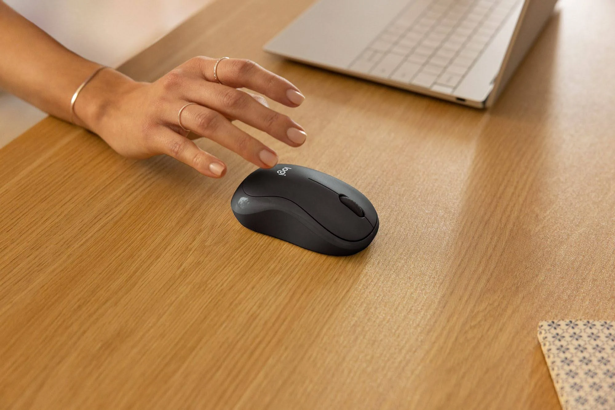 Une main tendue vers une souris d'ordinateur noire sur un bureau en bois. Un ordinateur portable est en arrière-plan.