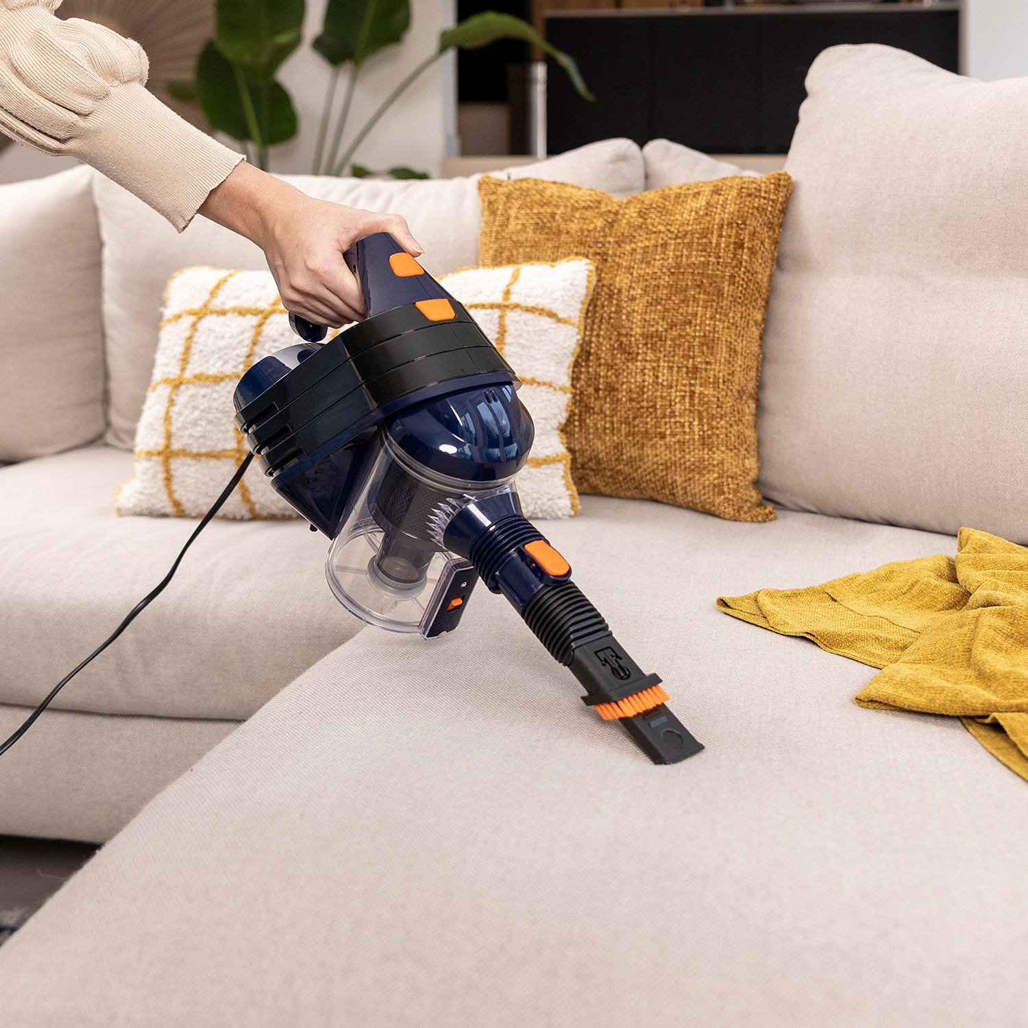 Una persona aspira un divano con un aspirapolvere portatile. L'aspirapolvere è blu navy e arancione. Il divano è beige.