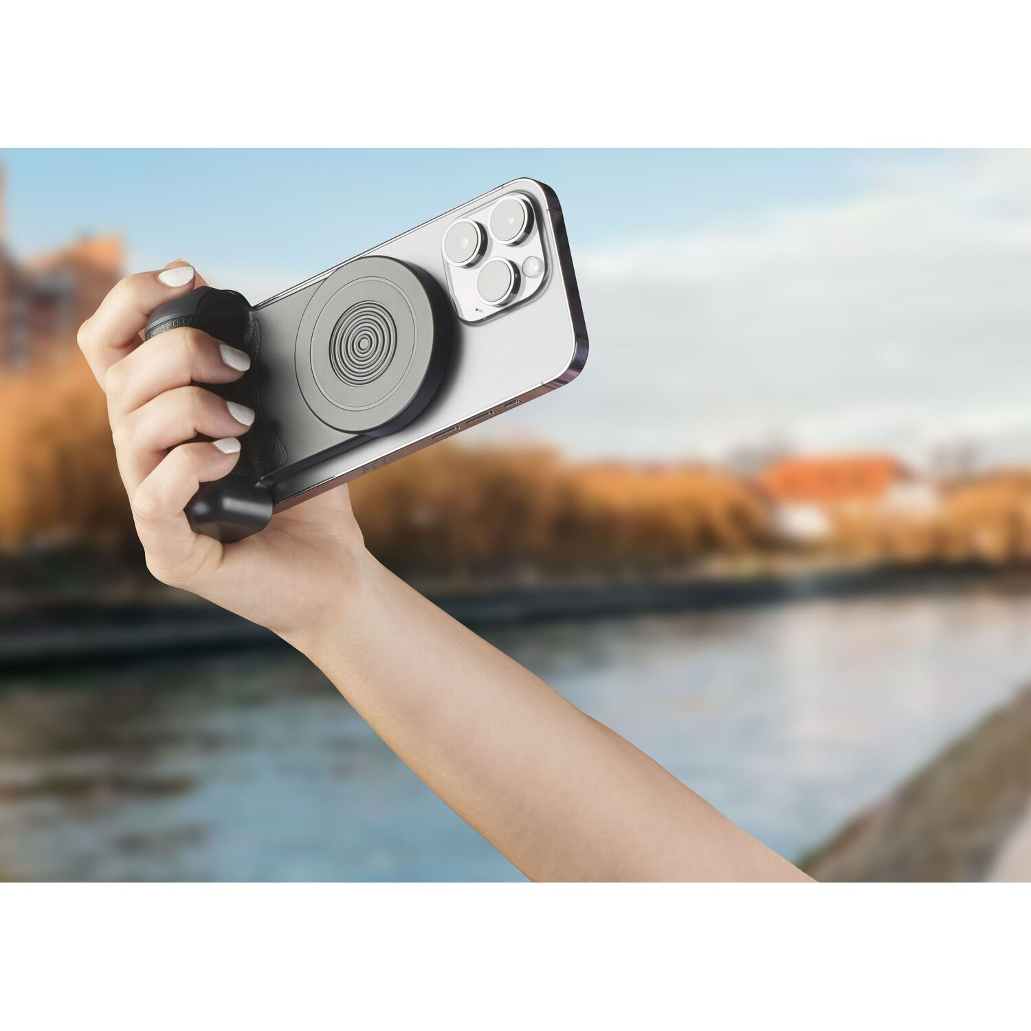 Hand holding a phone with a camera grip attached, over water with trees in the background.