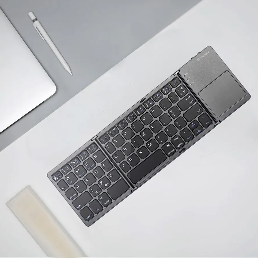 Faltbare, graue Tastatur mit Touchpad, Stift und Laptop teilweise auf grauer Oberfläche sichtbar.