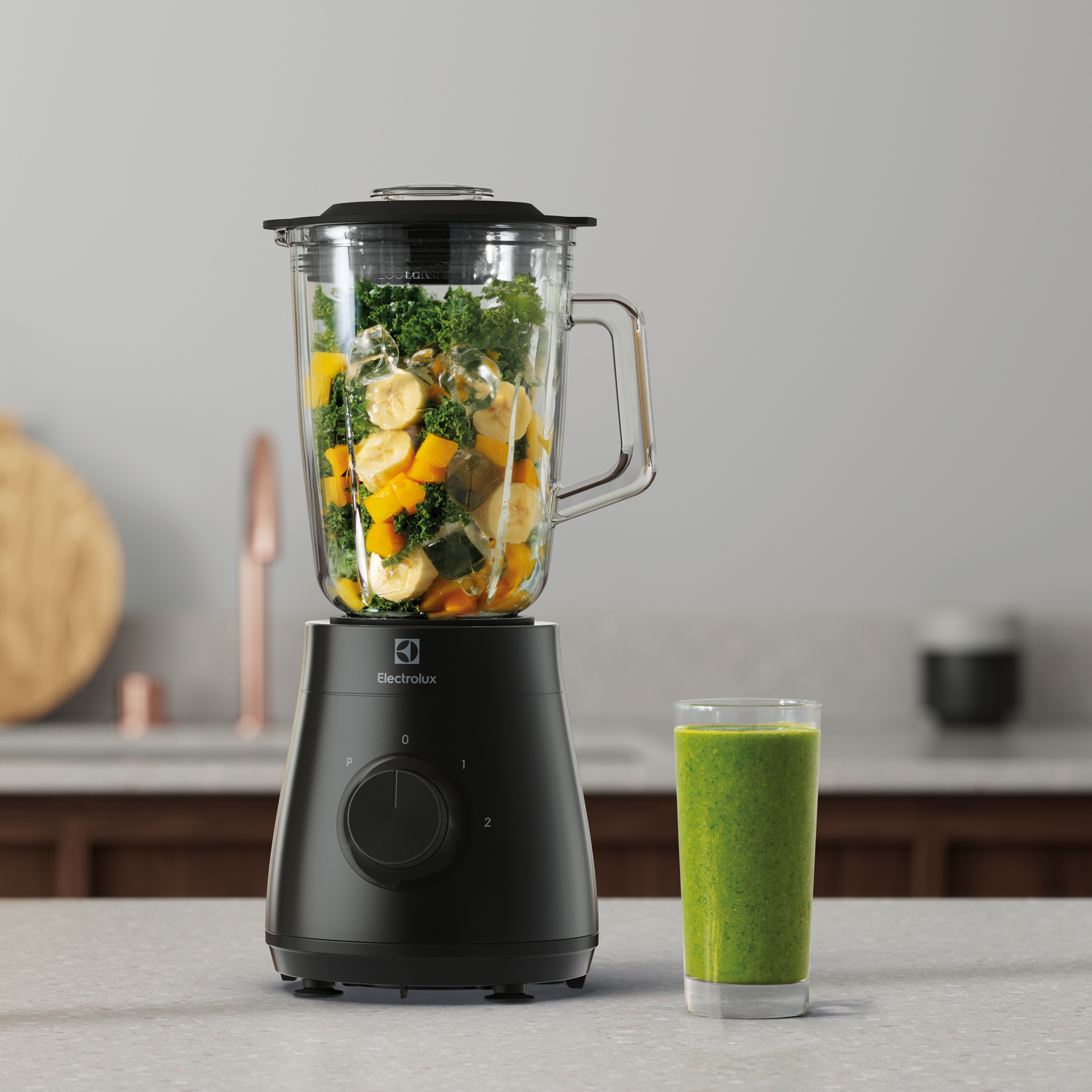 Un frullatore nero con ingredienti all'interno, un bicchiere di frullato verde, su un piano cucina. Frutta e cavolo riccio nel frullatore.