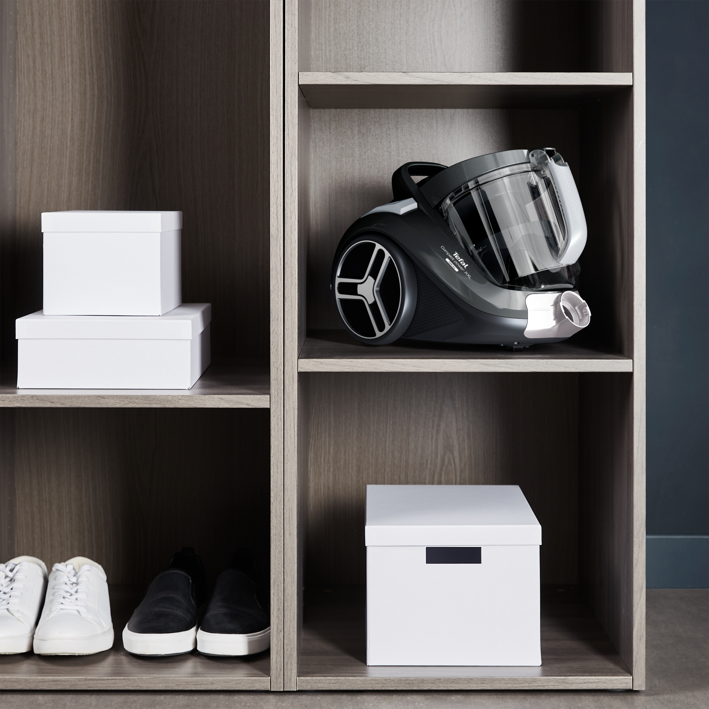 Drewniana szafka z odkurzaczem, pudełkami i butami. Ciemne drewniane półki. Szary odkurzacz, białe pudełka i buty.