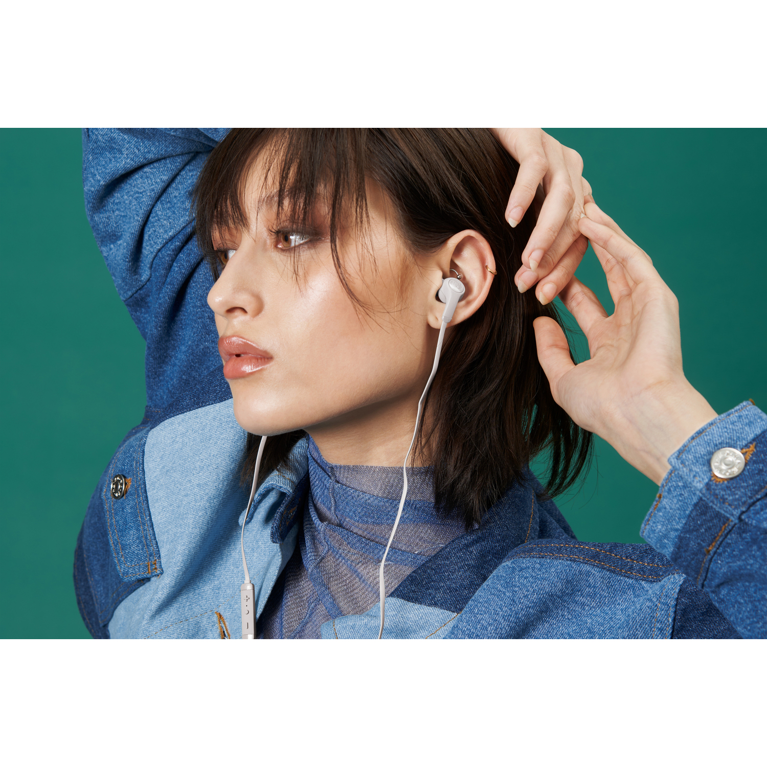 A woman wearing white earbuds and a denim jacket, looking to the left. Green background.
