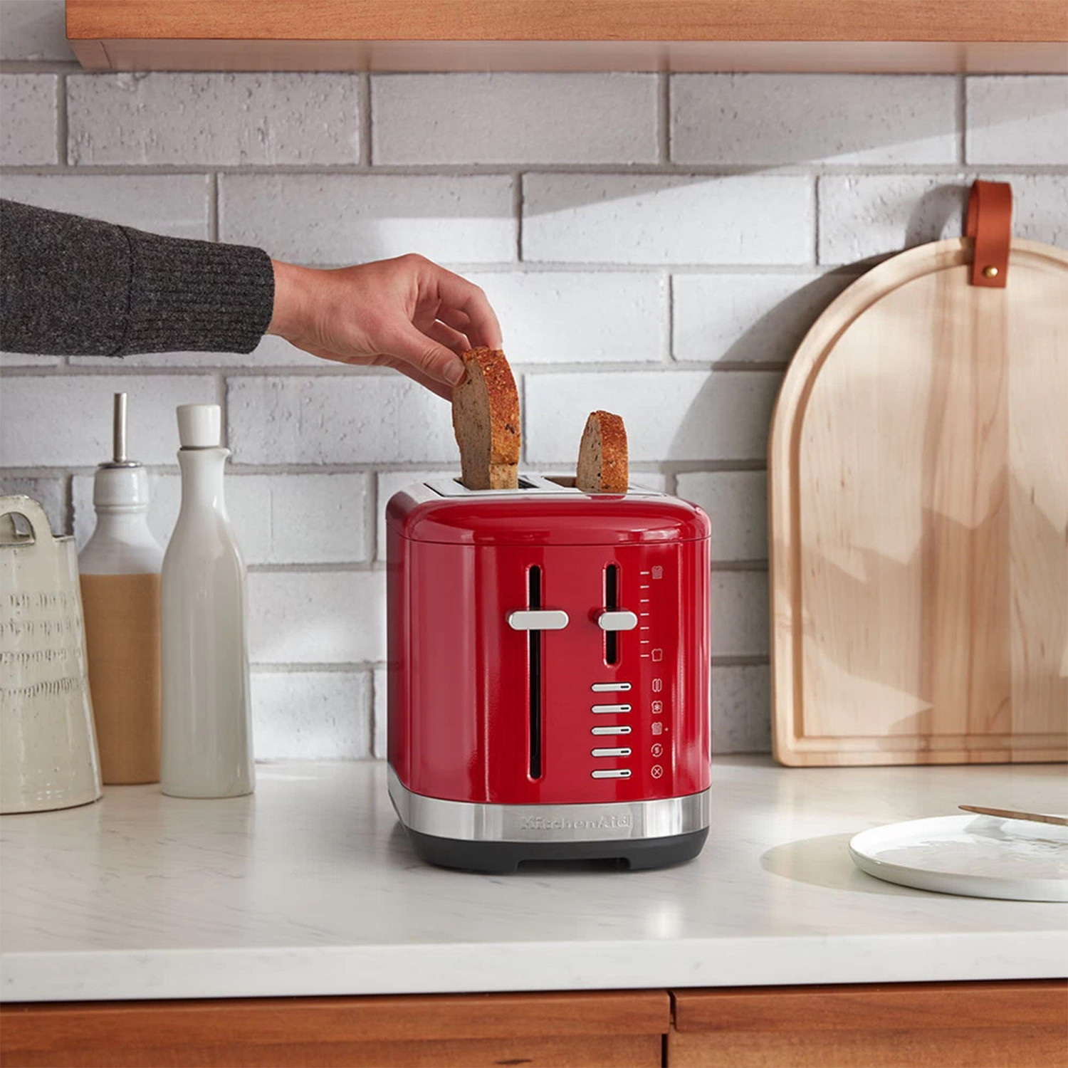 Un tostapane KitchenAid rosso con due fette di pane che vengono inserite. Dietro, un tagliere in legno.