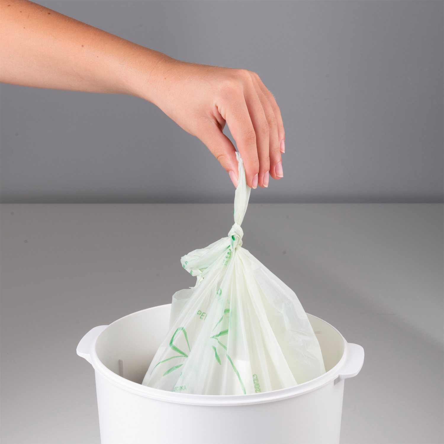 Una mano lega un sacco verde sopra un bidone della spazzatura bianco.