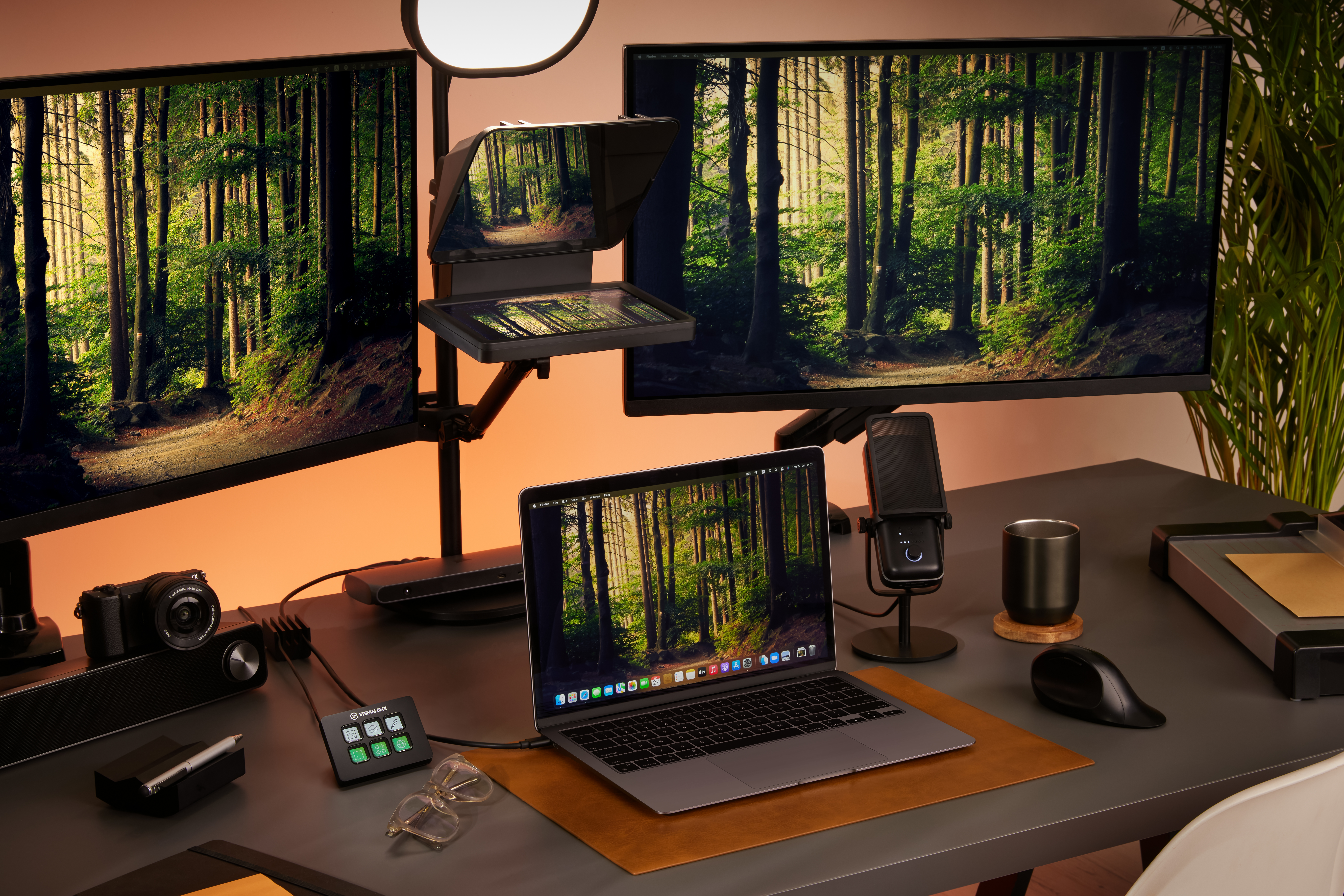 Ein Schreibtisch mit zwei Monitoren, die eine Waldszene zeigen, einem Laptop und Gadgets.