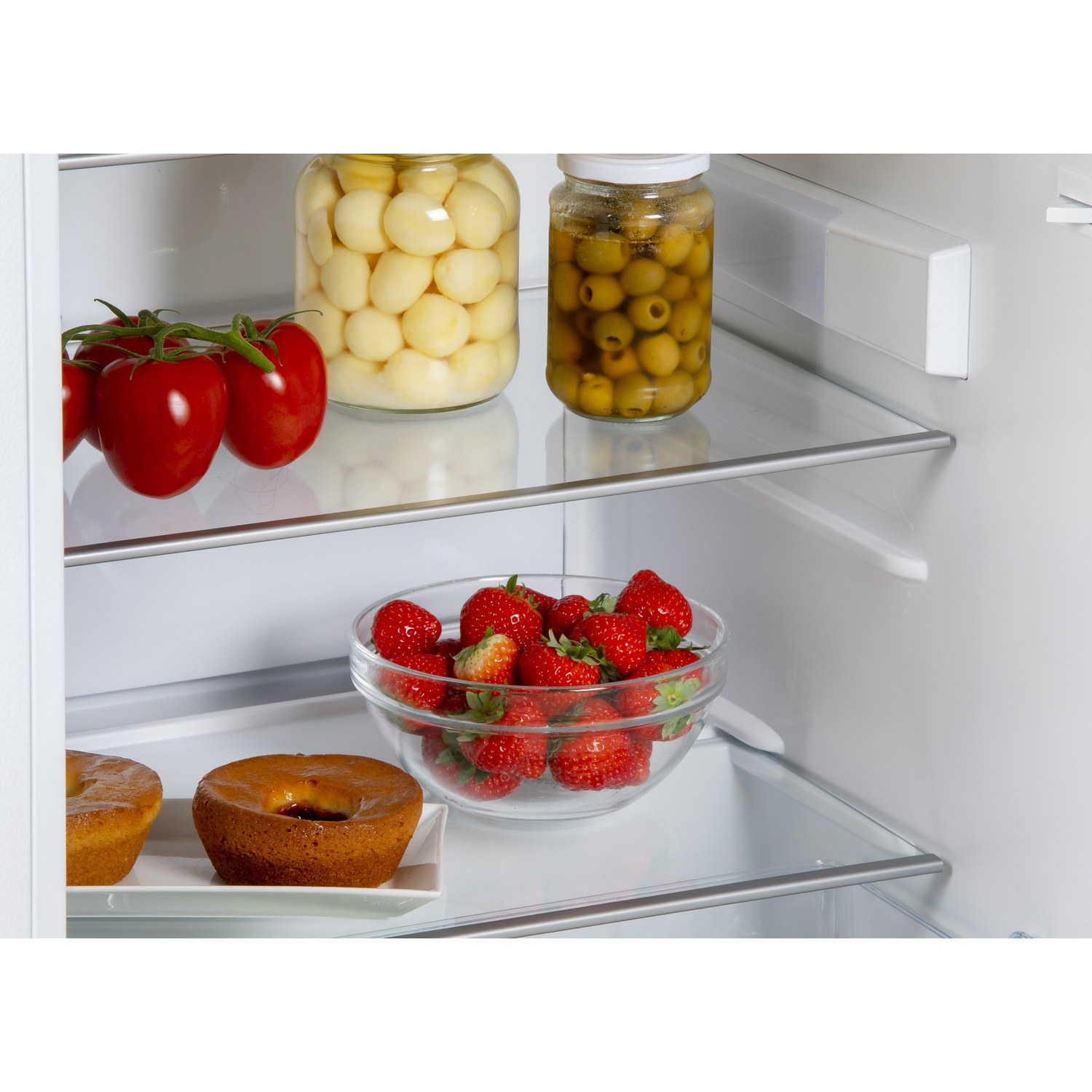 Inside a fridge: jars of pickled onions and olives, tomatoes, strawberries, and cakes.