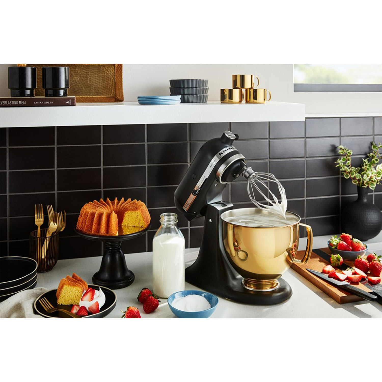 Un mixer KitchenAid nero e oro con una torta, fragole e altri ingredienti per la cottura nelle vicinanze.