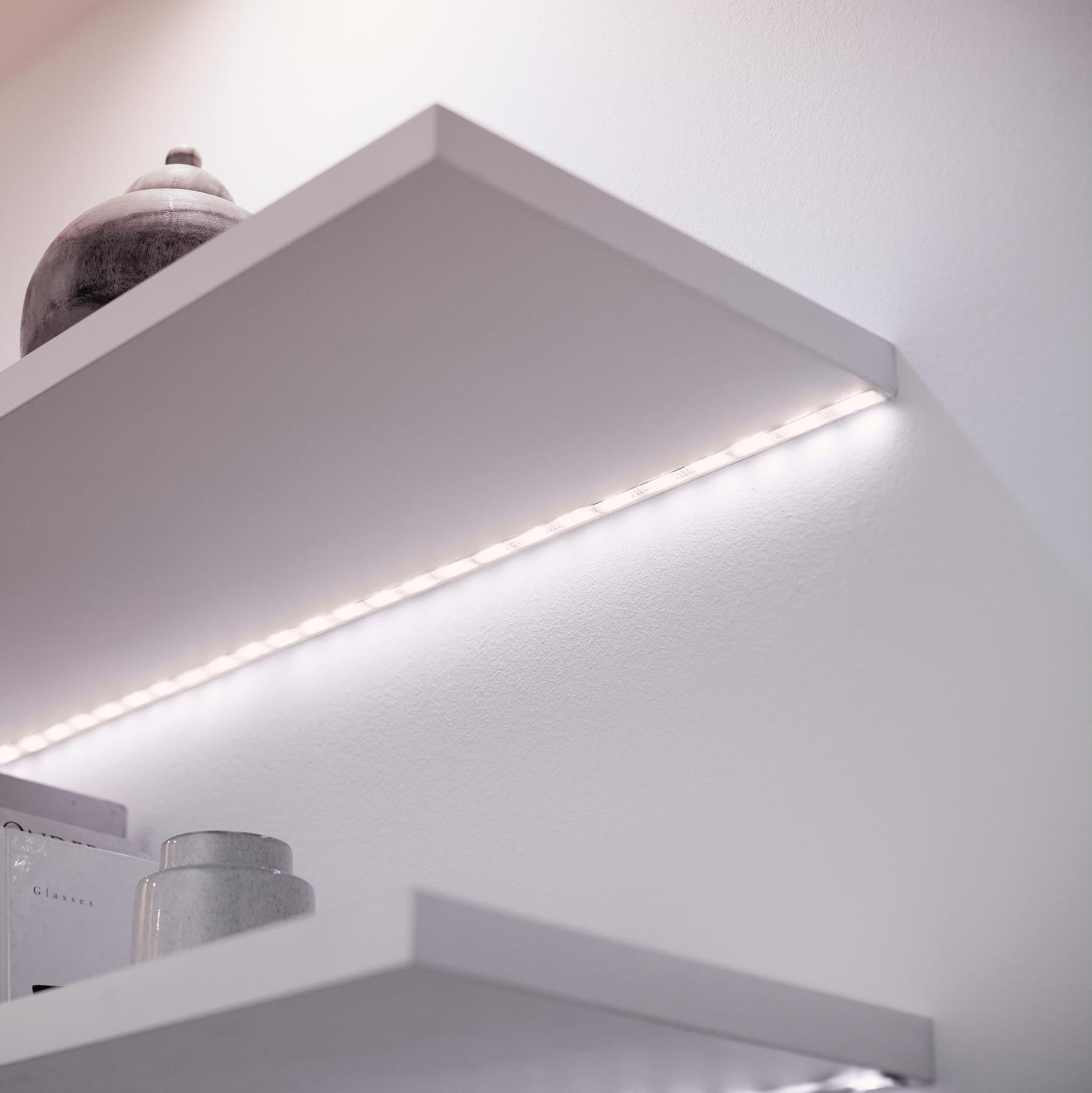 Mensole bianche con strisce luminose a LED sulla parete. Ci sono vasi e libri.