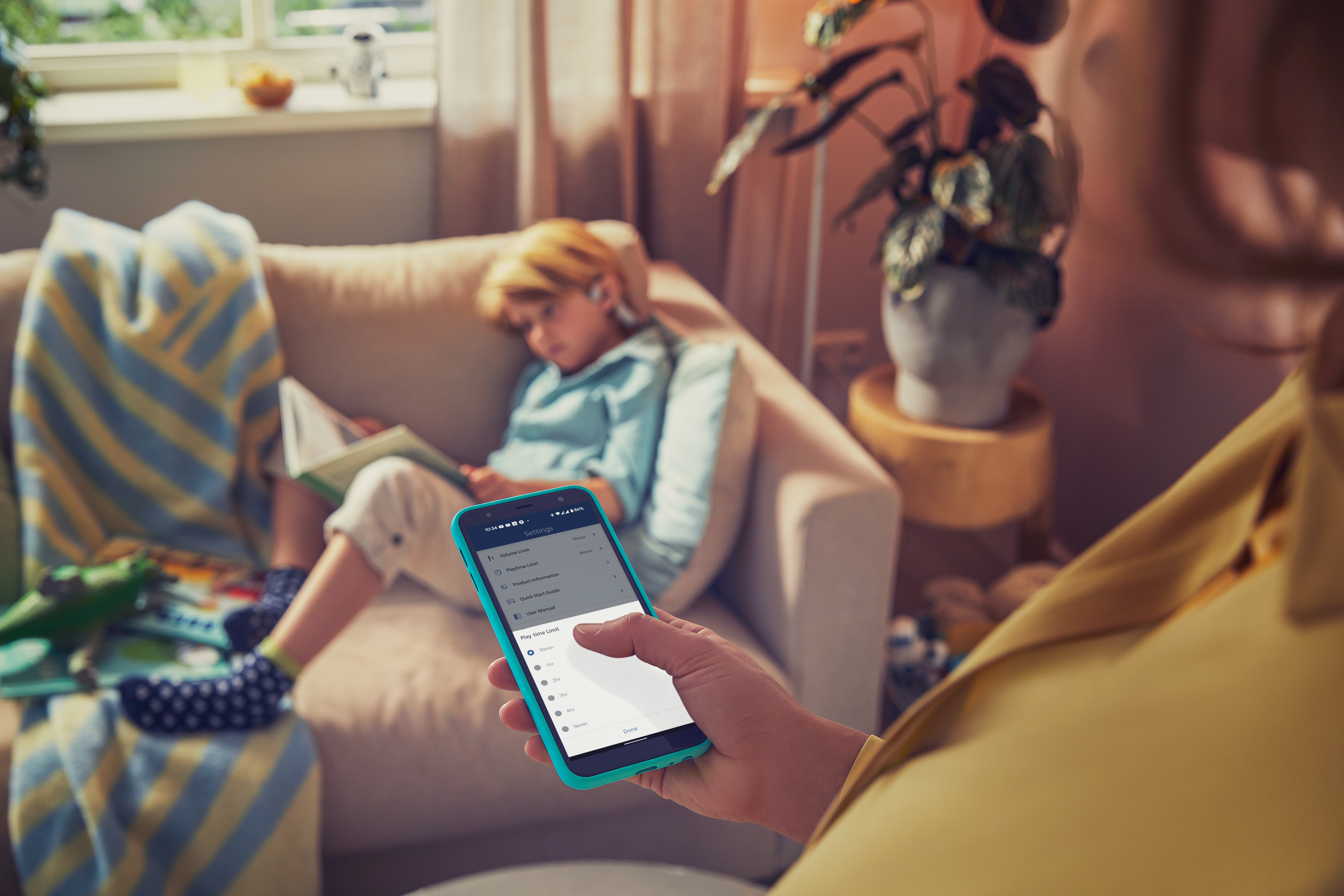 Person hält türkisfarbenes Smartphone mit Einstellungen, Kind liest auf dem Sofa.