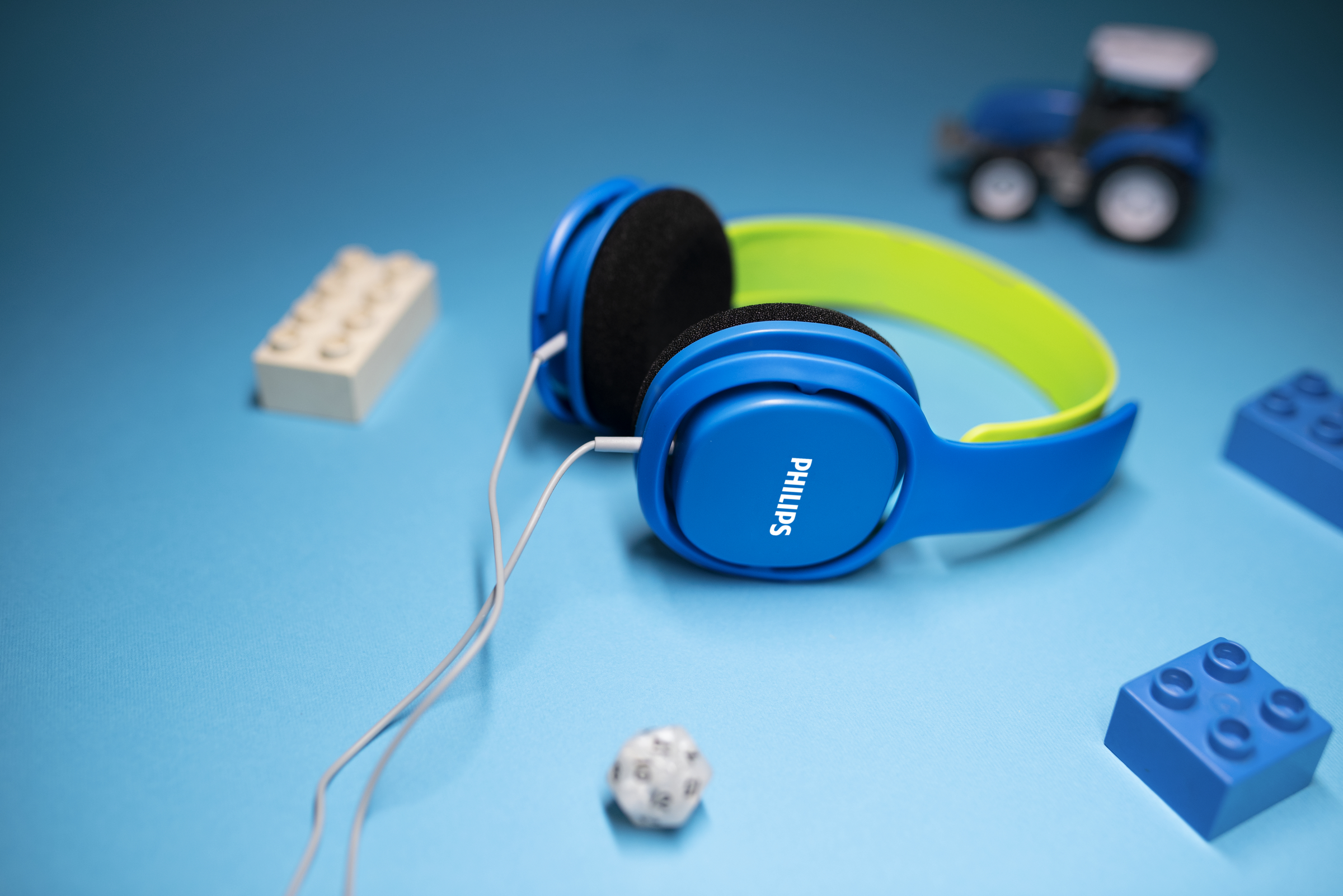 Blaue Kopfhörer mit grünem Band. LEGO-Blöcke und ein Spielzeugtraktor liegen auf blauer Oberfläche.