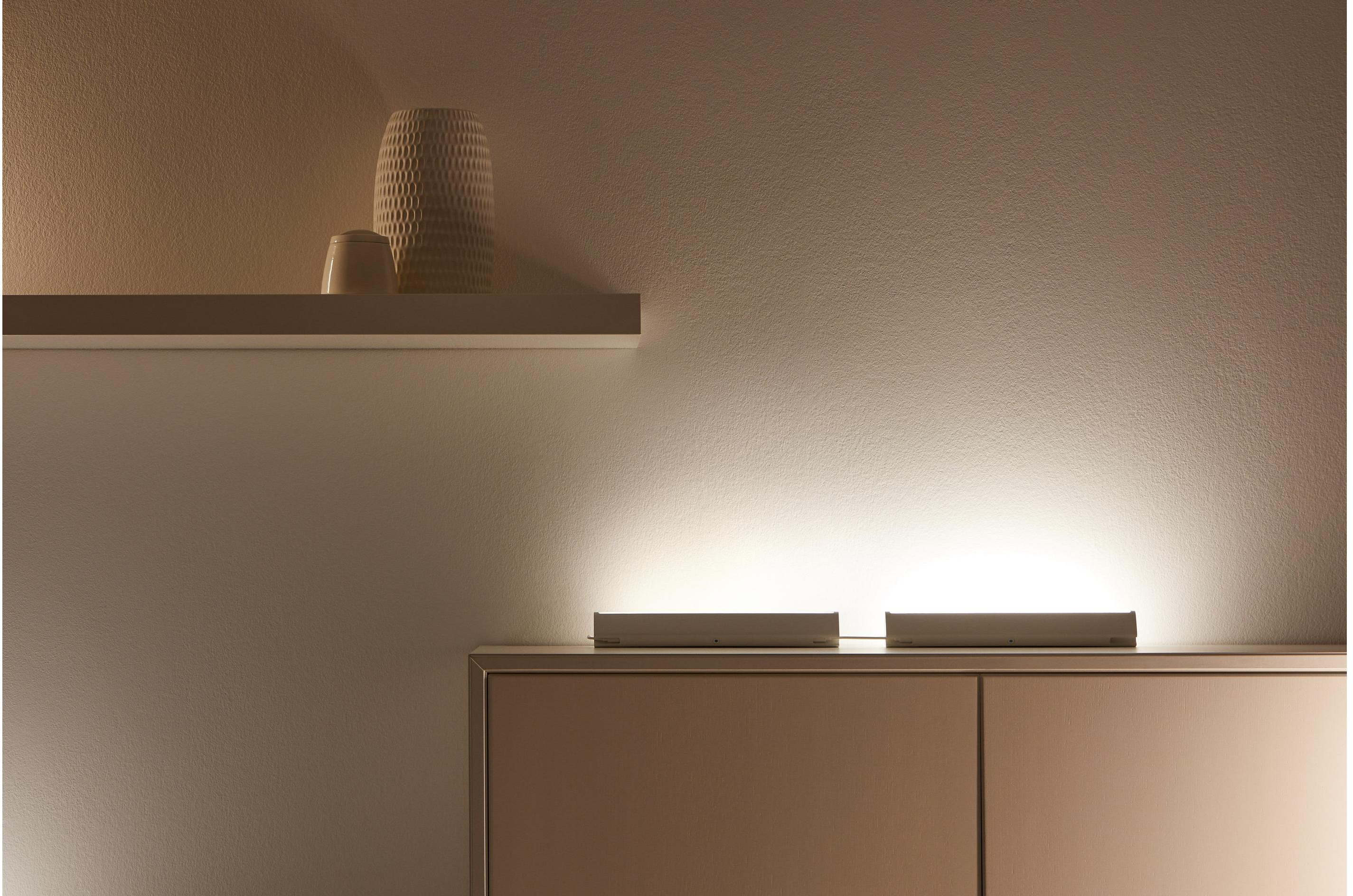 Interno di una stanza con due barre luminose bianche su un mobile e una mensola bianca con decorazioni.