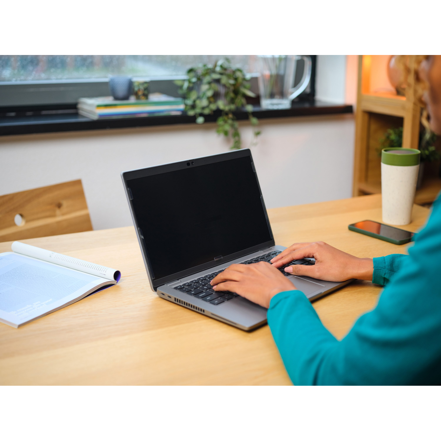 Persoon typt op een laptop aan een houten tafel. Een tijdschrift, een telefoon en een beker zijn zichtbaar. Groene plant en raam op de achtergrond.