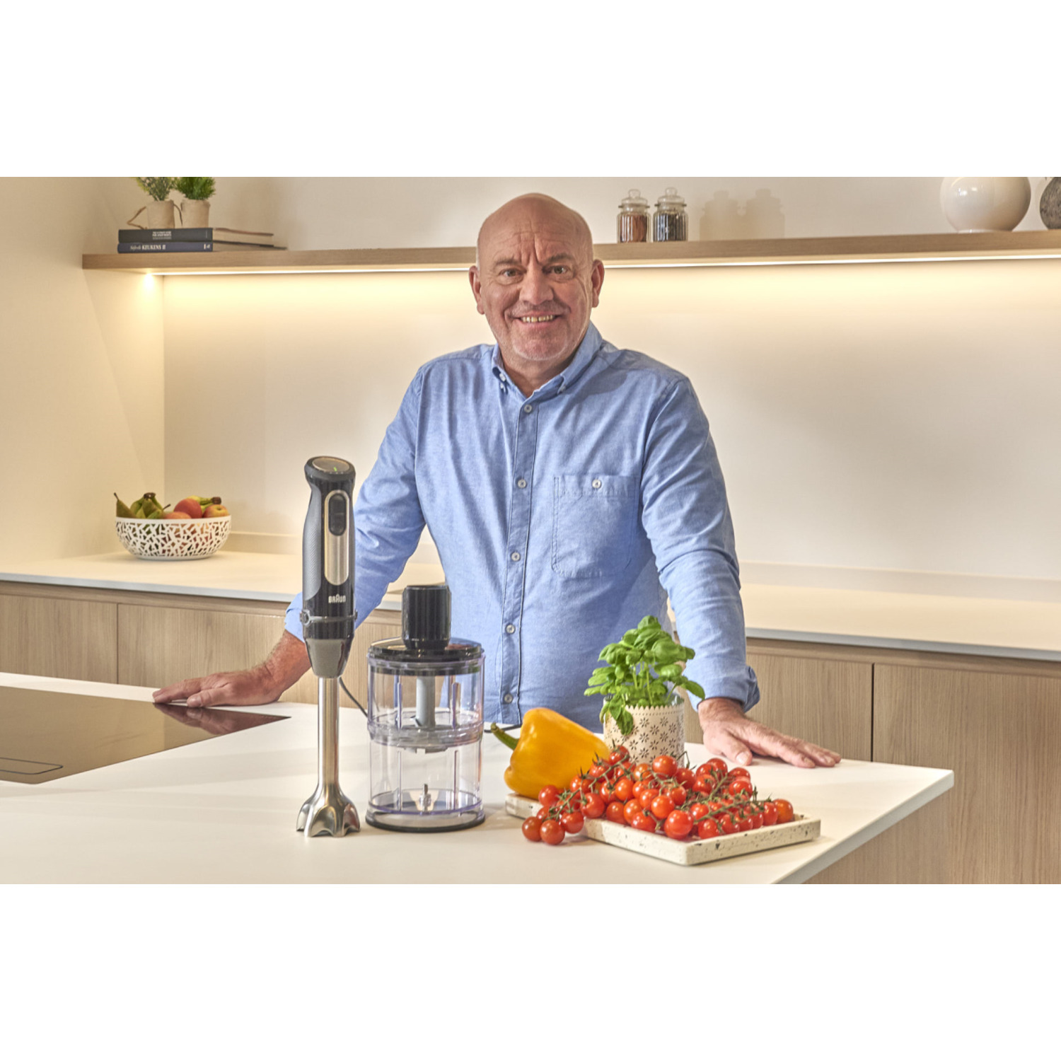 Man in a blue shirt stands at a white counter with a blender and vegetables.