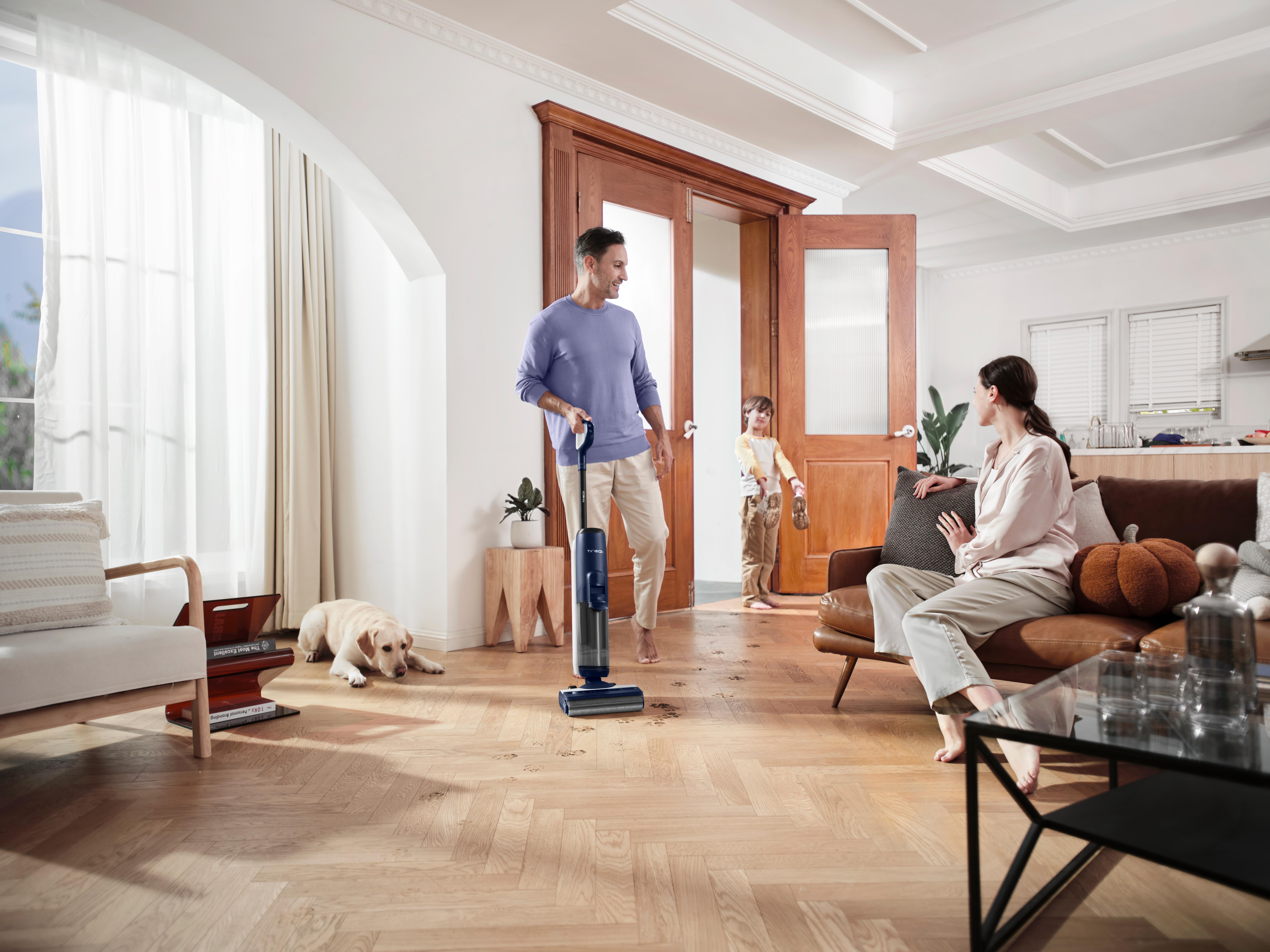 Eine Familie in einem Wohnzimmer. Ein Mann saugt einen Boden. Ein Hund und eine Frau sind auf dem Sofa.