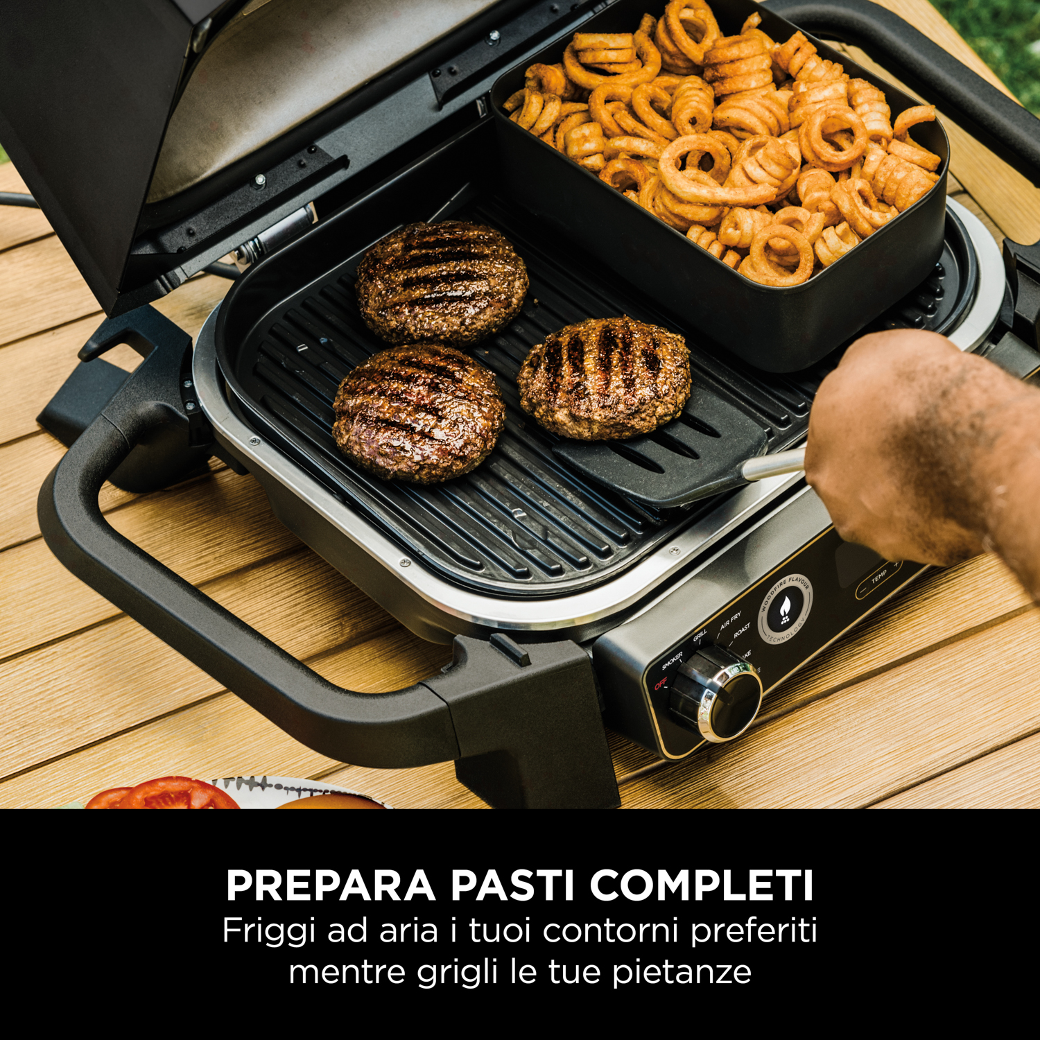 Griglia con hamburger e patatine fritte. Una mano usa una spatola. La griglia è su un tavolo di legno.