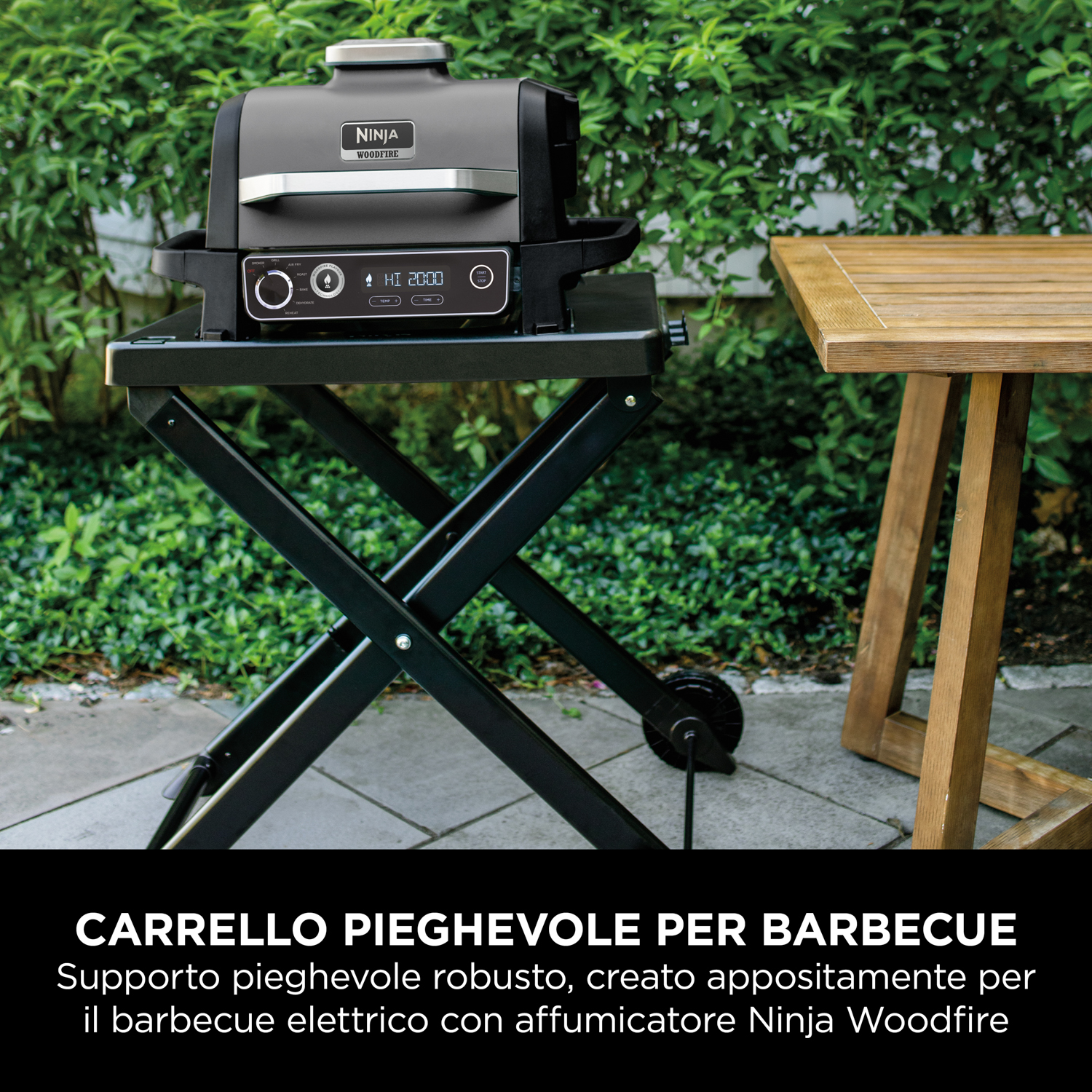 Griglia Ninja nera su un supporto pieghevole nero, vicino a un tavolo di legno. Arbusti verdi sullo sfondo.