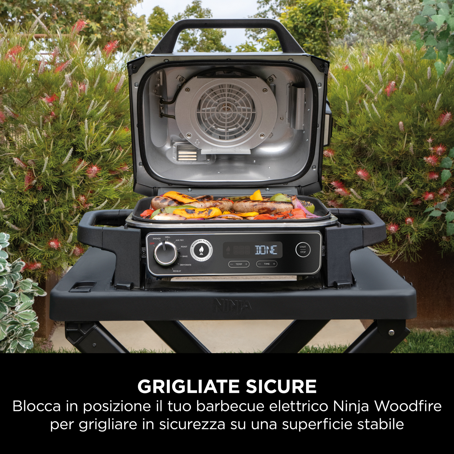 Griglia elettrica Ninja Woodfire con cibo dentro. Tavolo nero, piante verdi sullo sfondo. Coperchio aperto.