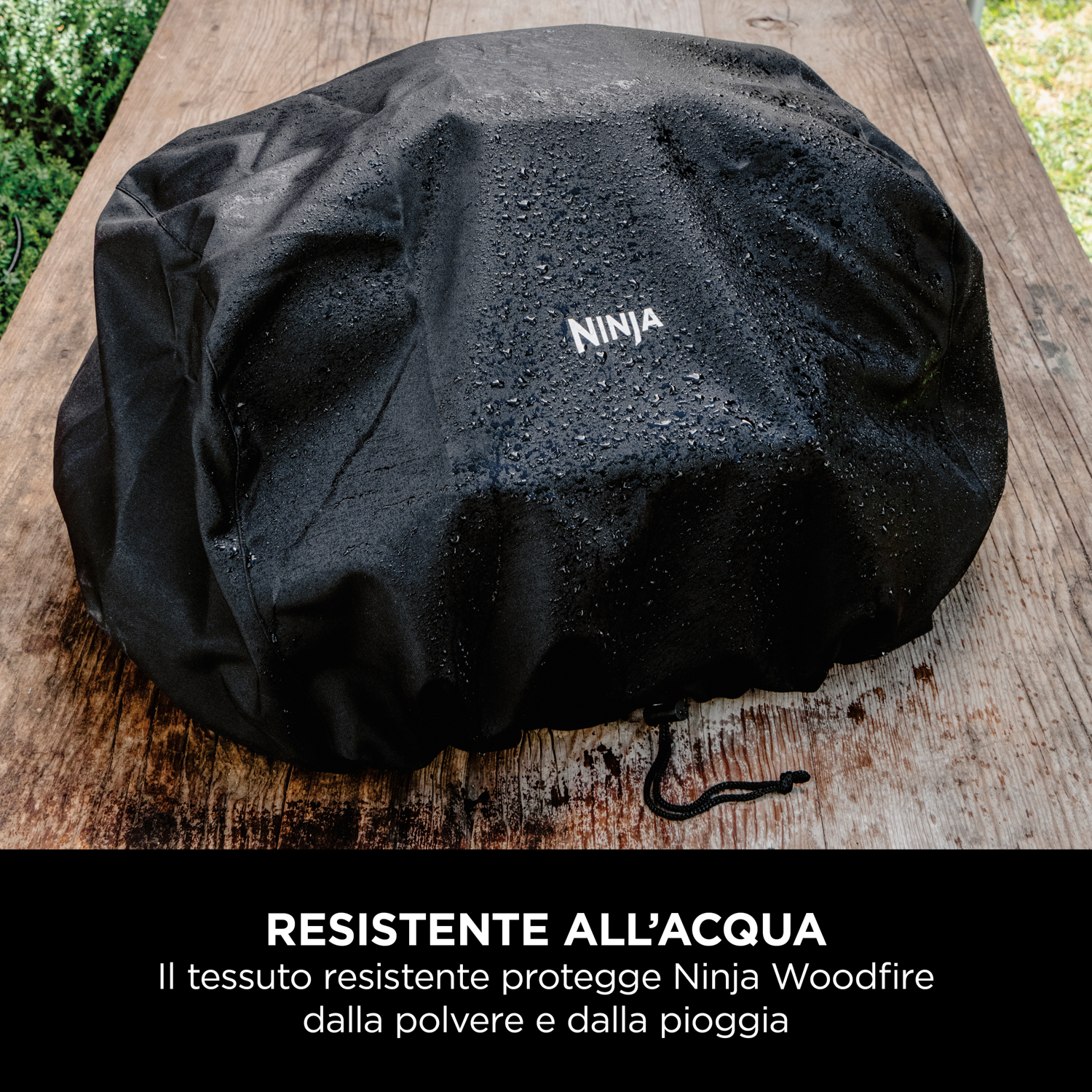 Copertura nera per grill Ninja Woodfire, goccioline d'acqua sopra, su un tavolo di legno.