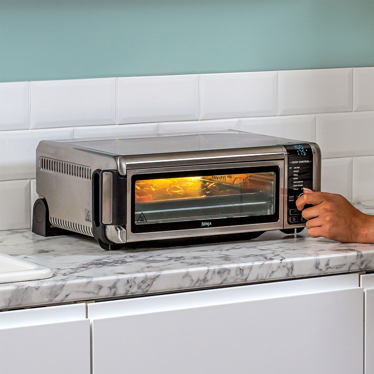 Un forno tostapane in acciaio inossidabile su un piano di lavoro con la mano di una persona che regola un quadrante. Cibo all'interno illuminato.