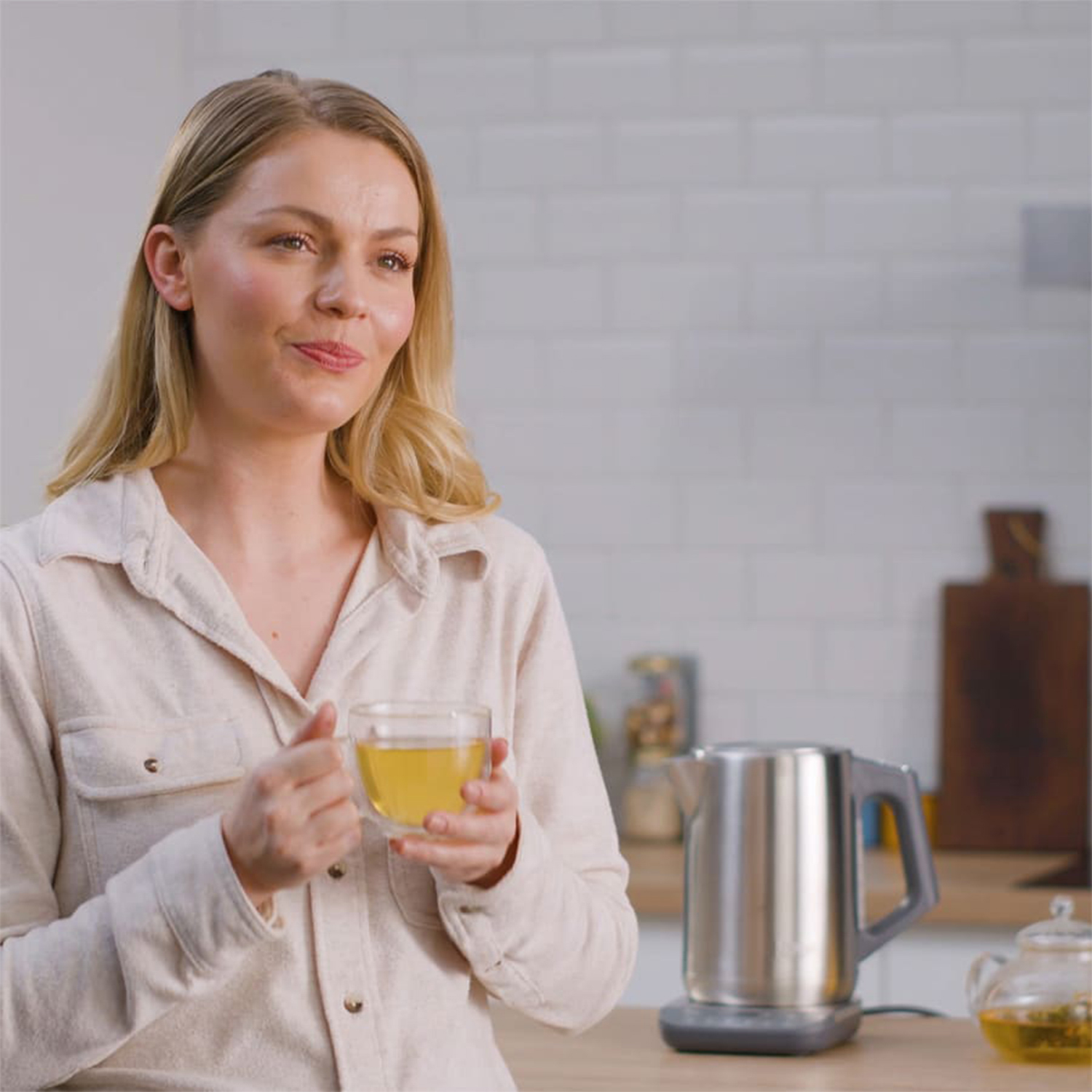 Donna bionda che tiene un bicchiere di tè, in cucina, con un bollitore.