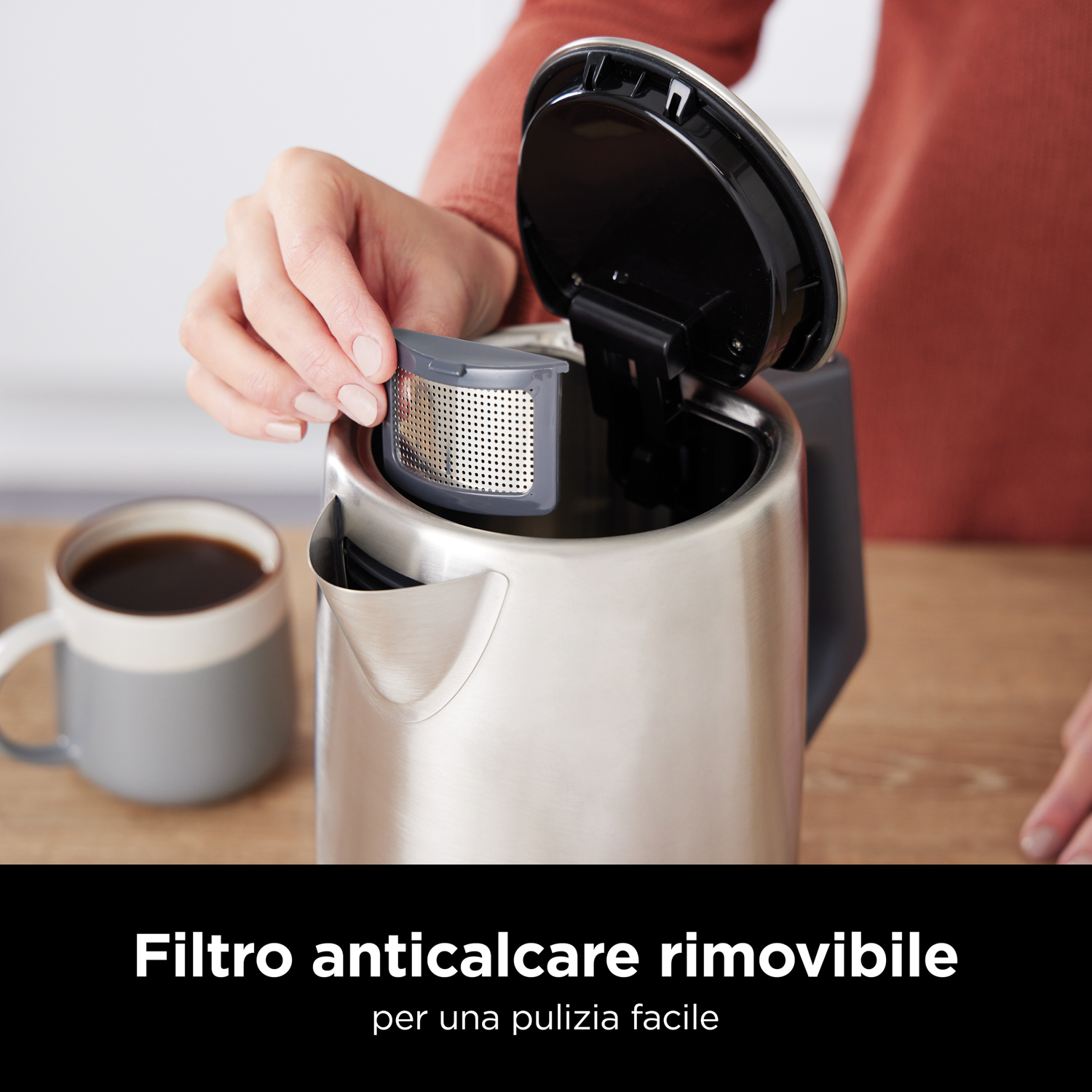 Una mano rimuove un filtro da un bollitore in acciaio inossidabile, tazza e caffè visibili.