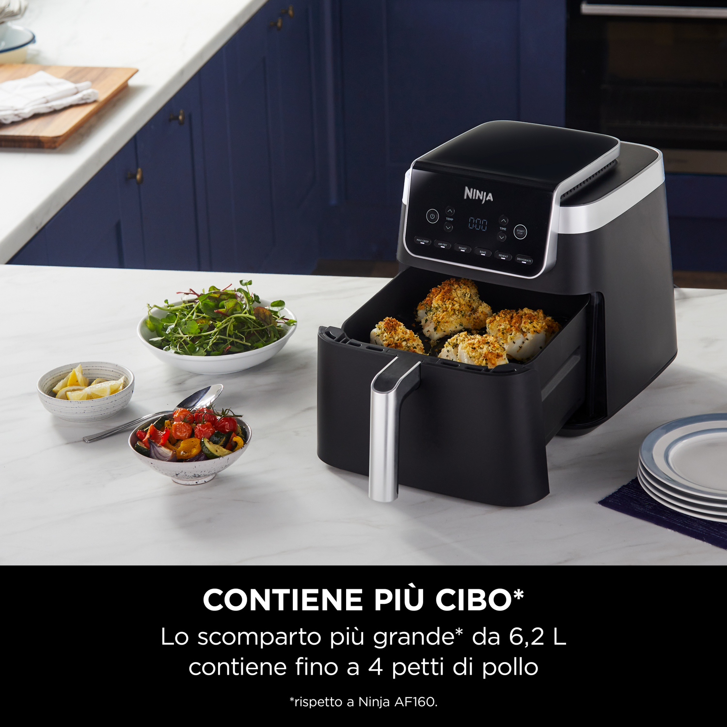 Friggitrice ad aria Ninja nera con cibo all'interno, ciotole con cibo accanto e un piano di lavoro bianco.
