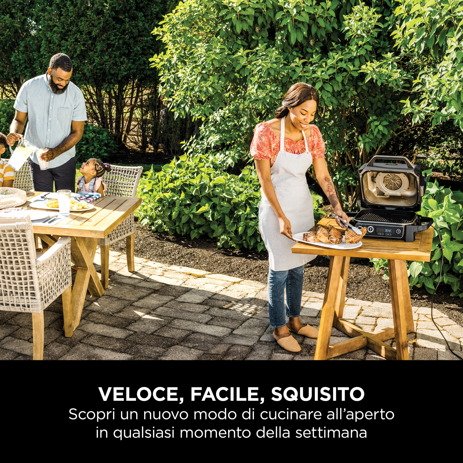 Una famiglia all'aperto, che cucina cibo su una griglia Ninja. Sono visibili un tavolo e delle sedie.
