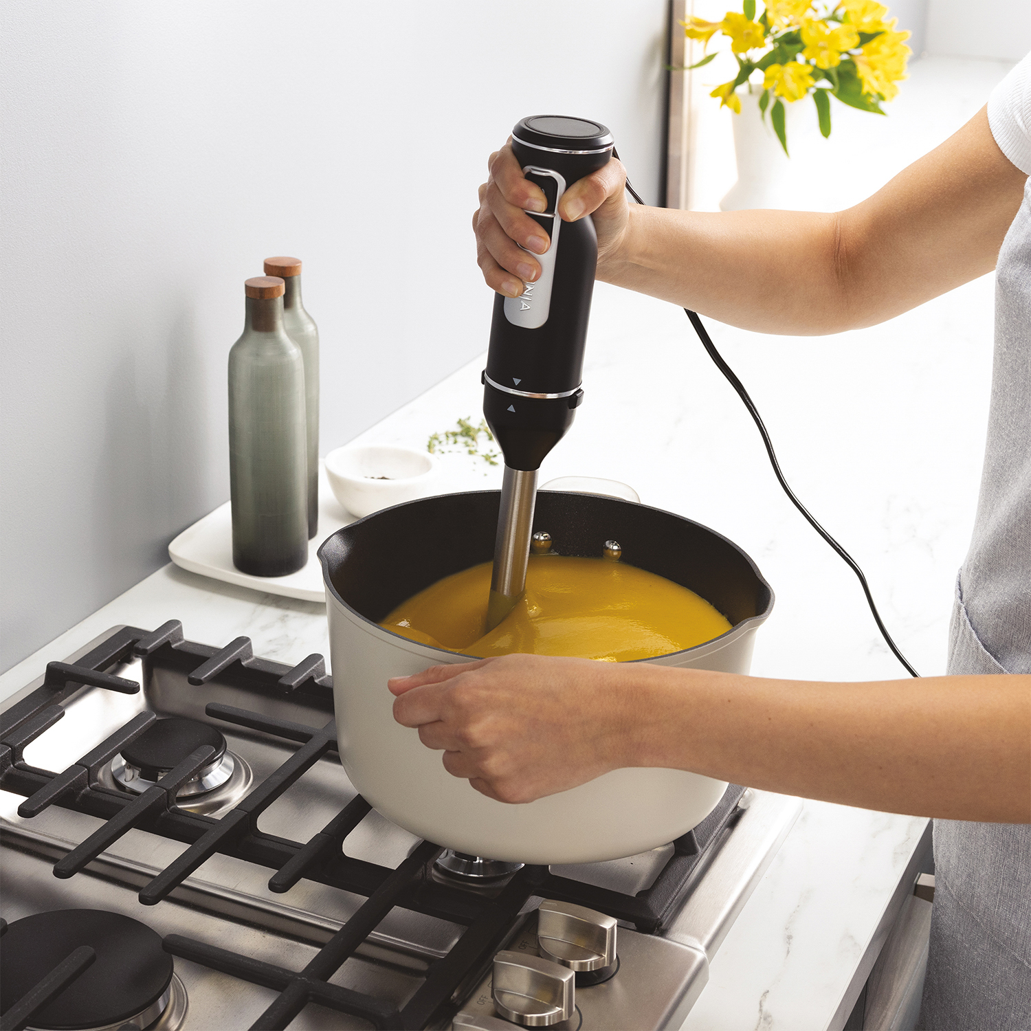 Persona che frulla la zuppa. Frullatore a immersione in una pentola. Zuppa gialla. Cucina grigia e bianca.