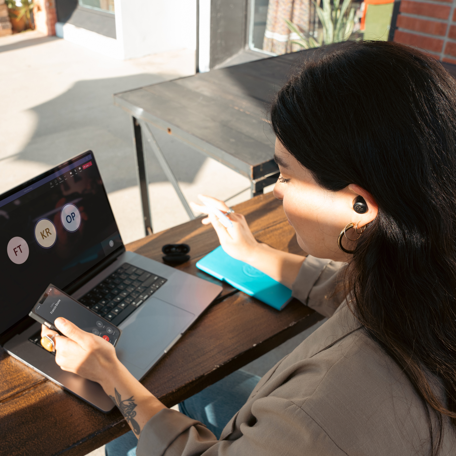 Woman using laptop and phone.  Earbuds in ear.  Blue charging pad on table.