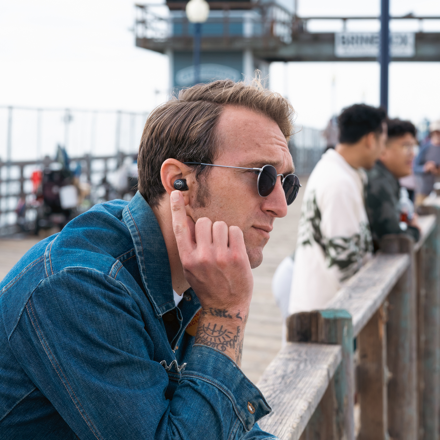 A man wearing sunglasses, with an earbud in one ear, and looking away from the camera. Pier is in background.