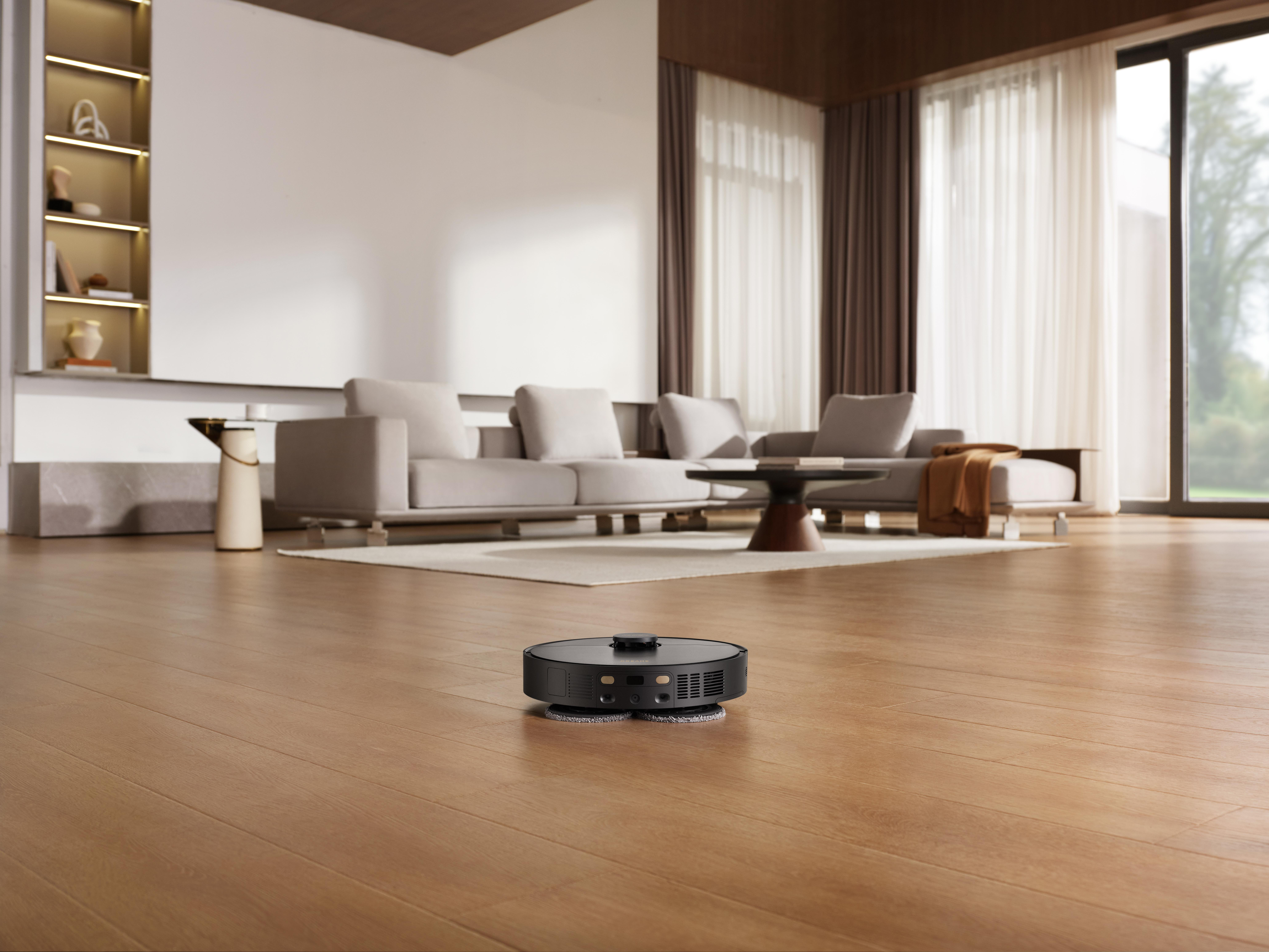 Ein schwarzer Roboter-Staubsauger reinigt einen braunen Holzboden. Im Hintergrund sind hellgraue Sofas zu sehen.