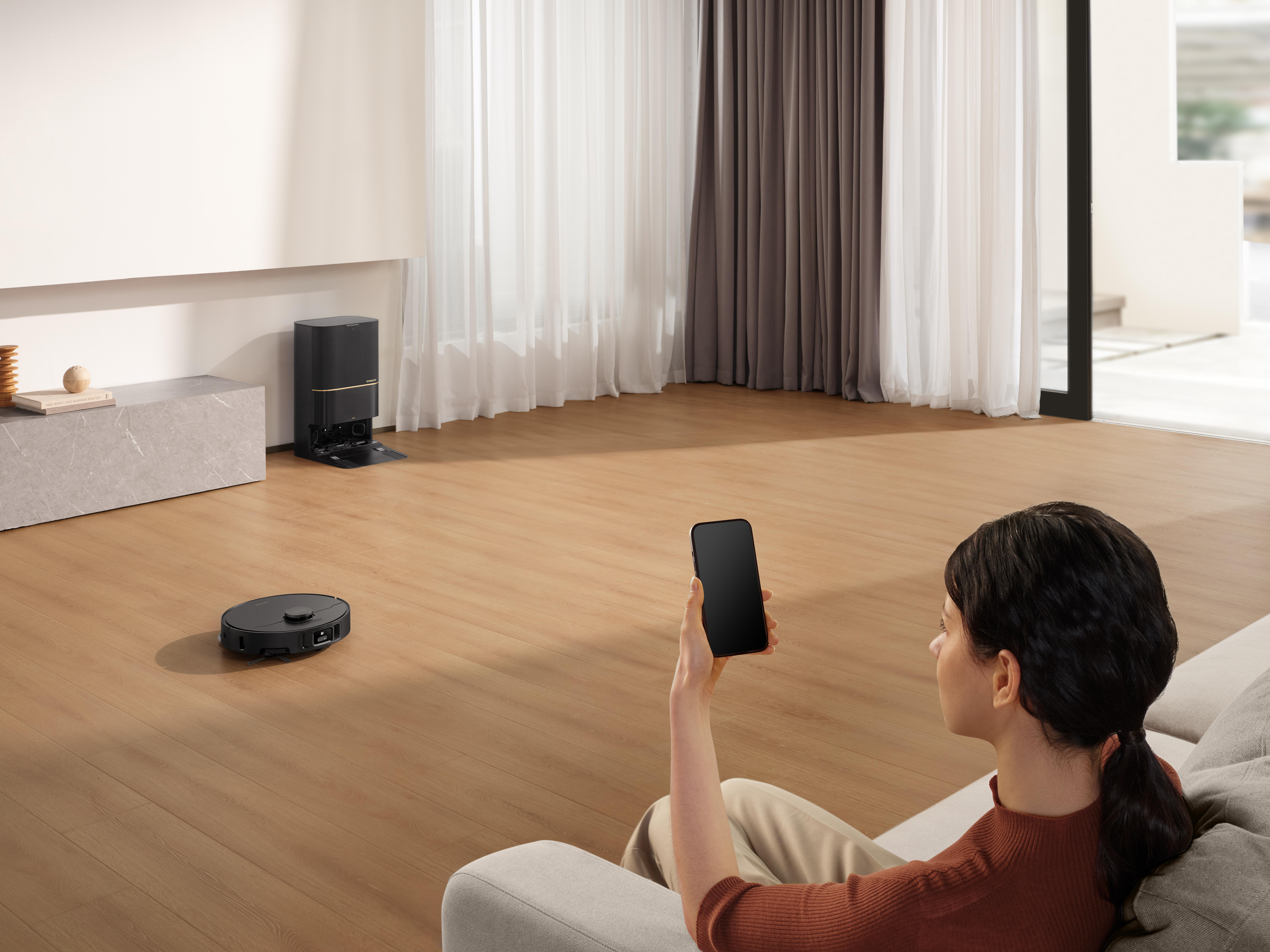 Frau steuert Roboter-Staubsauger per Telefon. Moderner Wohnraum im Hintergrund.