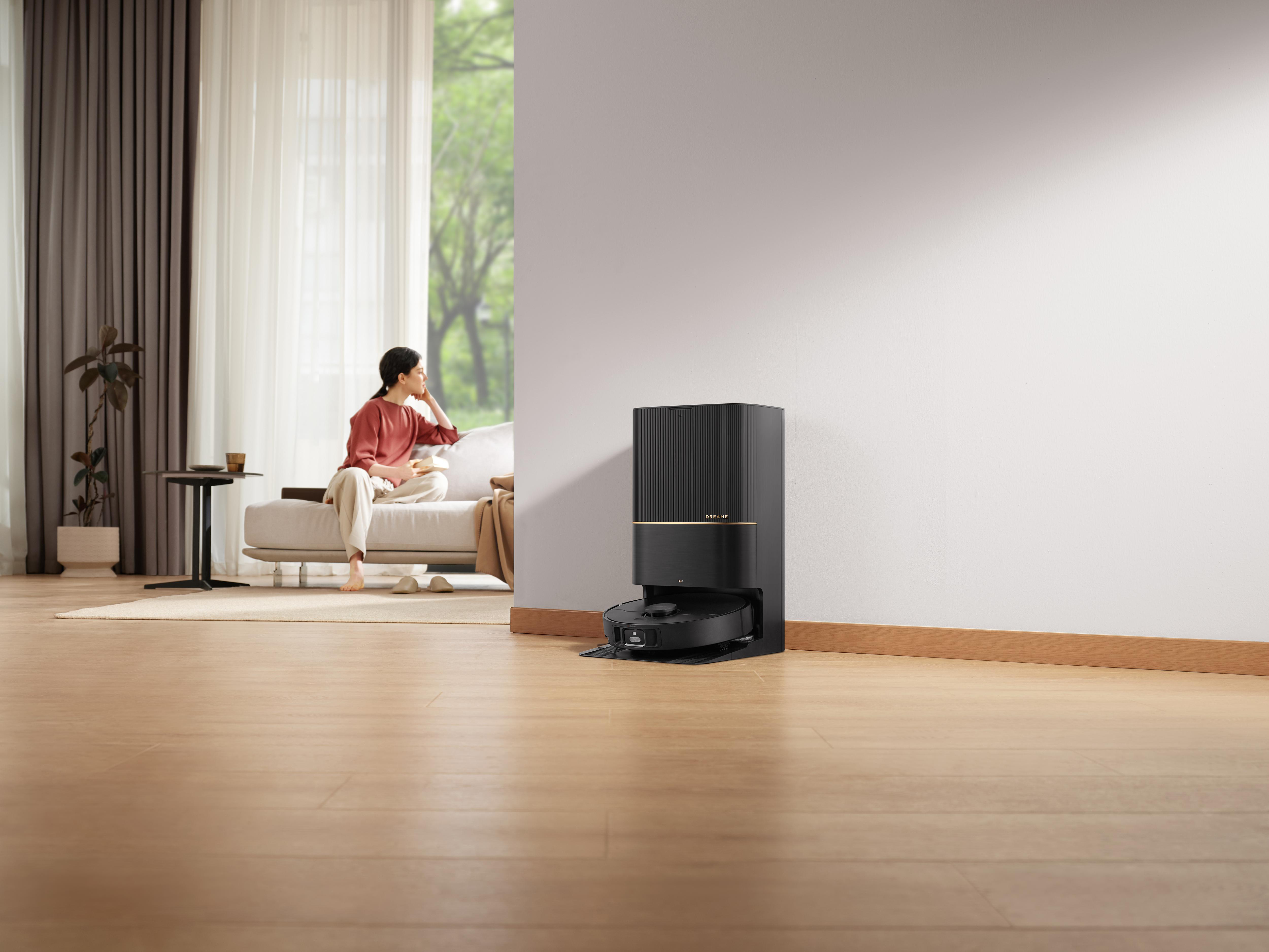 Eine Frau sitzt auf einem Sofa in einem Raum. Ein Roboter-Staubsauger reinigt den Boden.