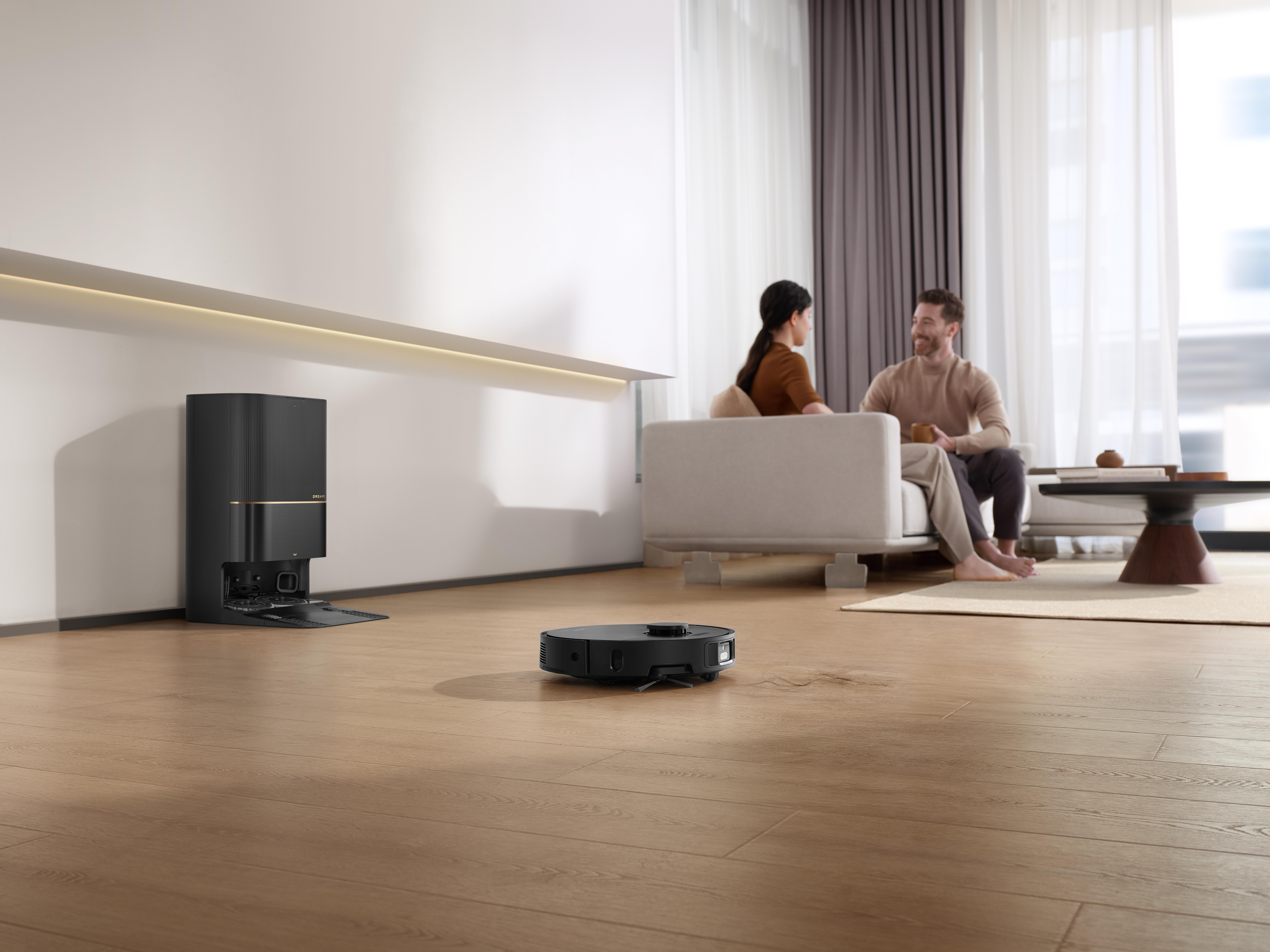 Ein Roboter-Staubsauger reinigt einen Holzboden. Personen sitzen im Hintergrund auf einem Sofa.