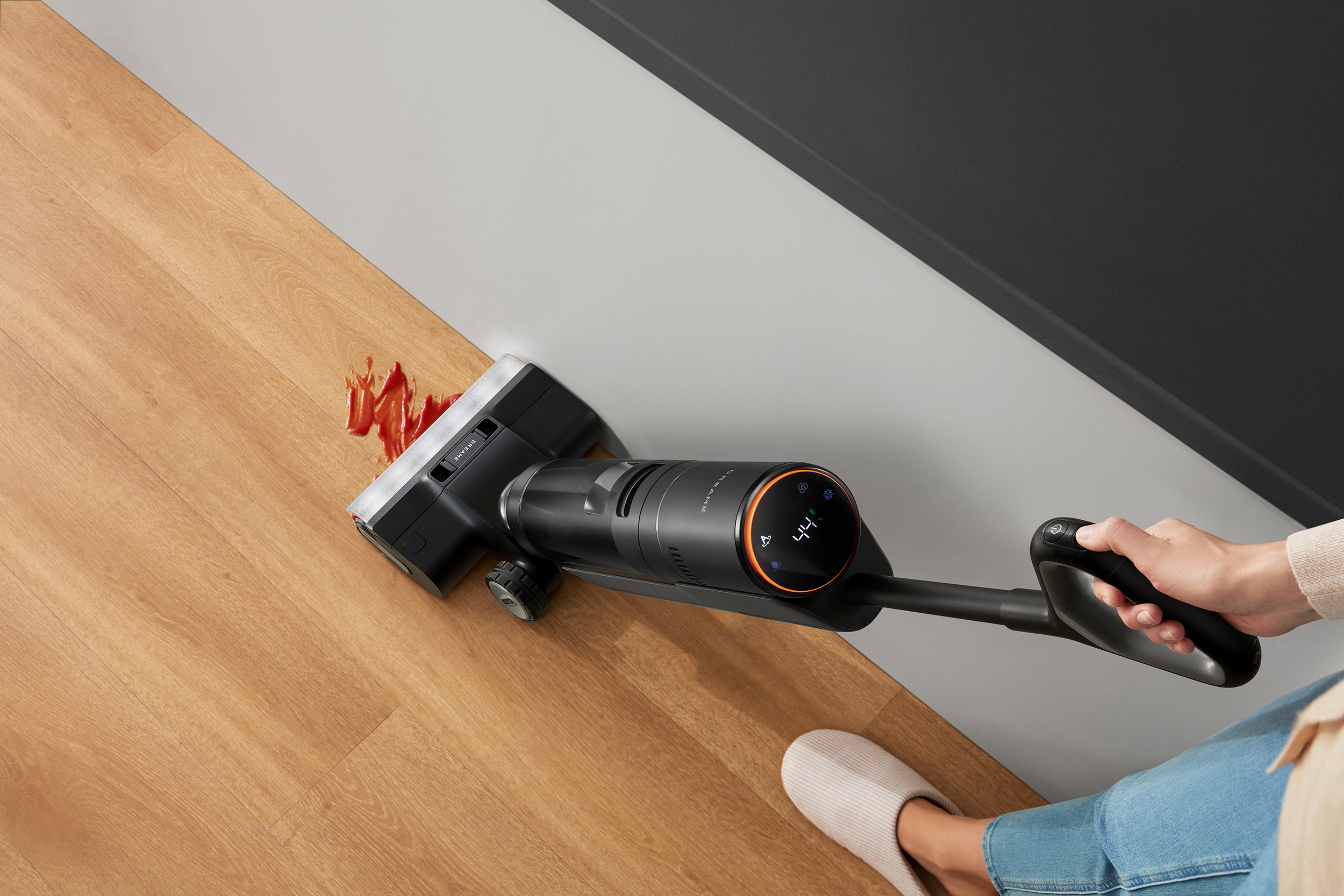 Eine Person reinigt einen Holzboden mit einem schwarz-orangen Staubsauger. Ein Fleck ist sichtbar.