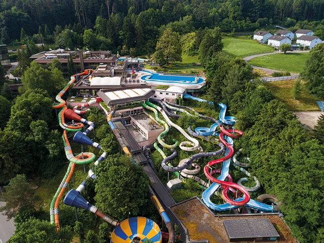 Vue aérienne d'un parc aquatique. Toboggans de différentes couleurs, piscines et bâtiments visibles.