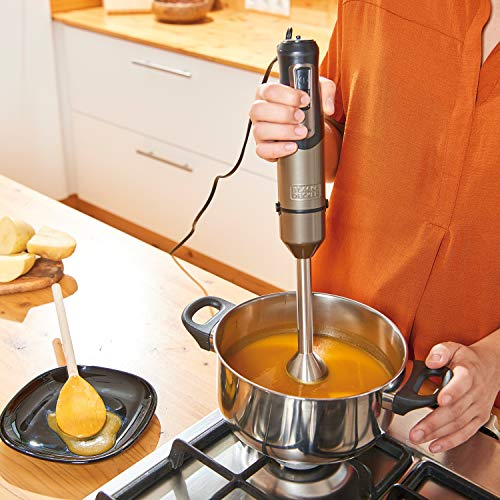 Osoba używająca blendera ręcznego w garnku z zupą i łyżką z olejem.