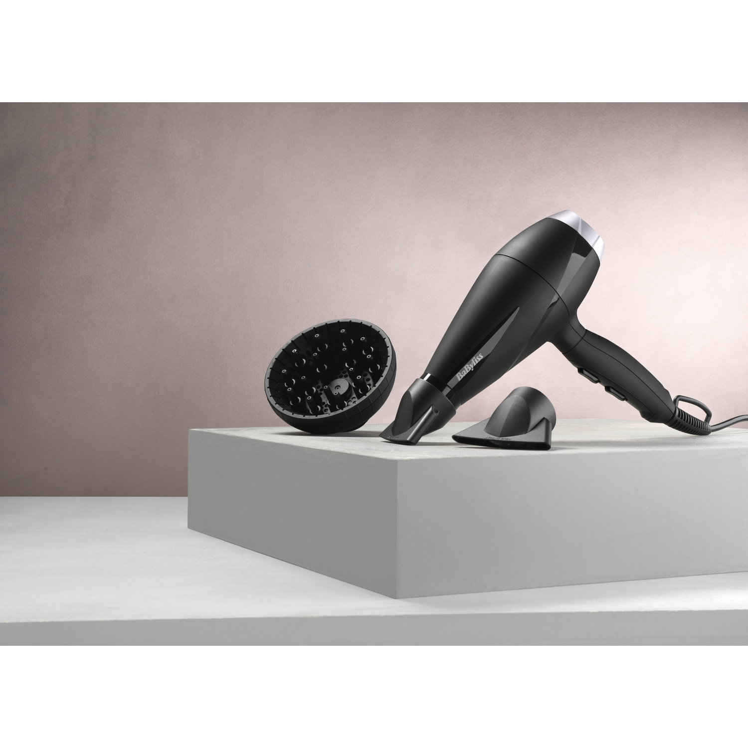 Black hair dryer, diffuser, and nozzle on a white platform and a pink background.