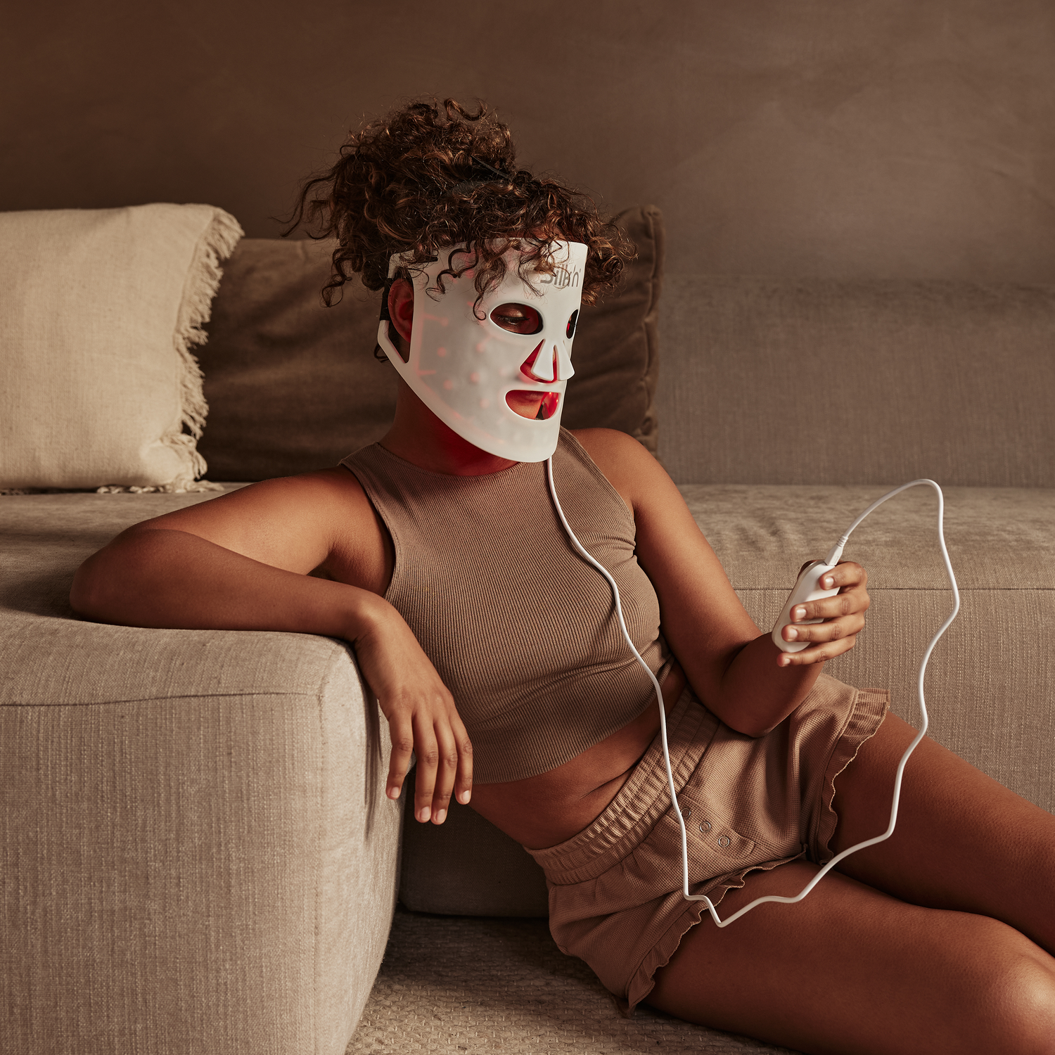 Vrouw met wit LED-masker, zittend op een bank, met controller.