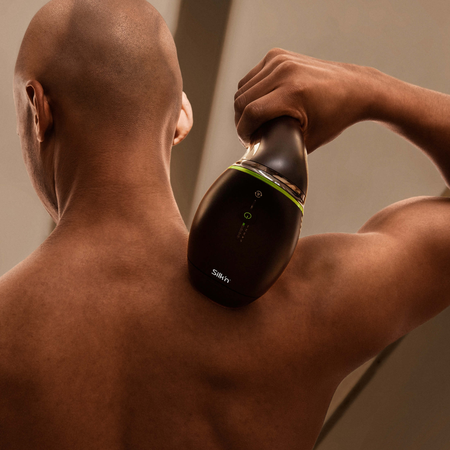 Bald man using a black massager on his shoulder. The device is held in his right hand.