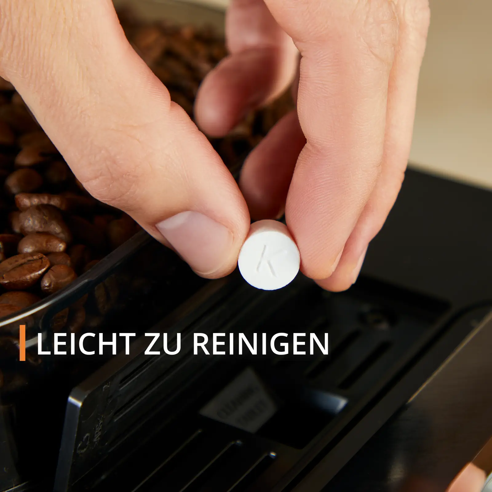 Eine Hand, die eine weiße Reinigungstablette mit dem Buchstaben 'K' vor Kaffeebohnen hält.