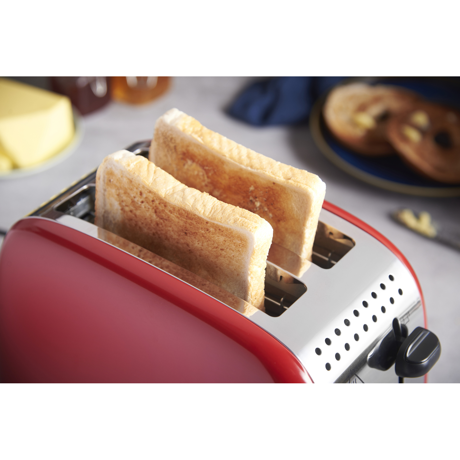 A red toaster with two slices of toasted bread. Butter, jam, and bread are in the background.