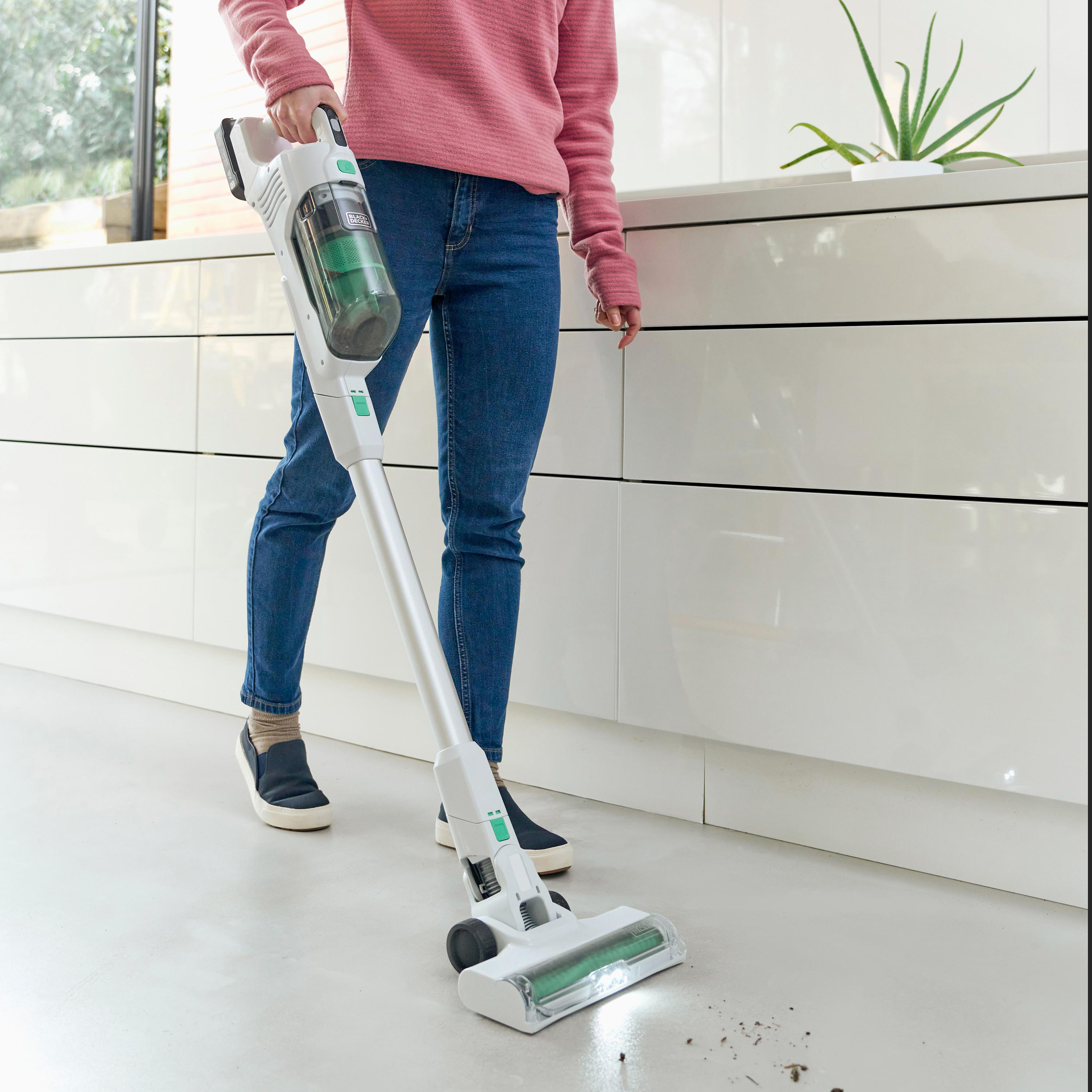 Person saugt Boden. Staubsauger ist weiß und grün, mit Pflanze im Hintergrund.