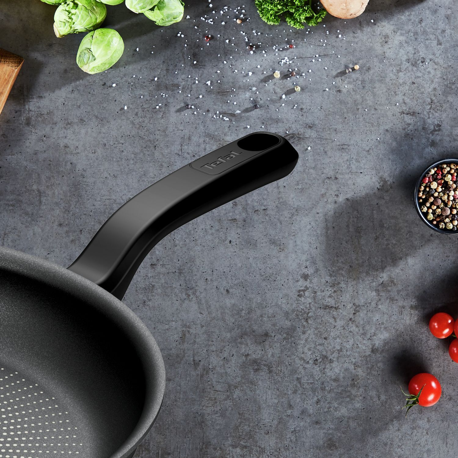 A black frying pan with a handle, surrounded by vegetables and spices on a gray surface.